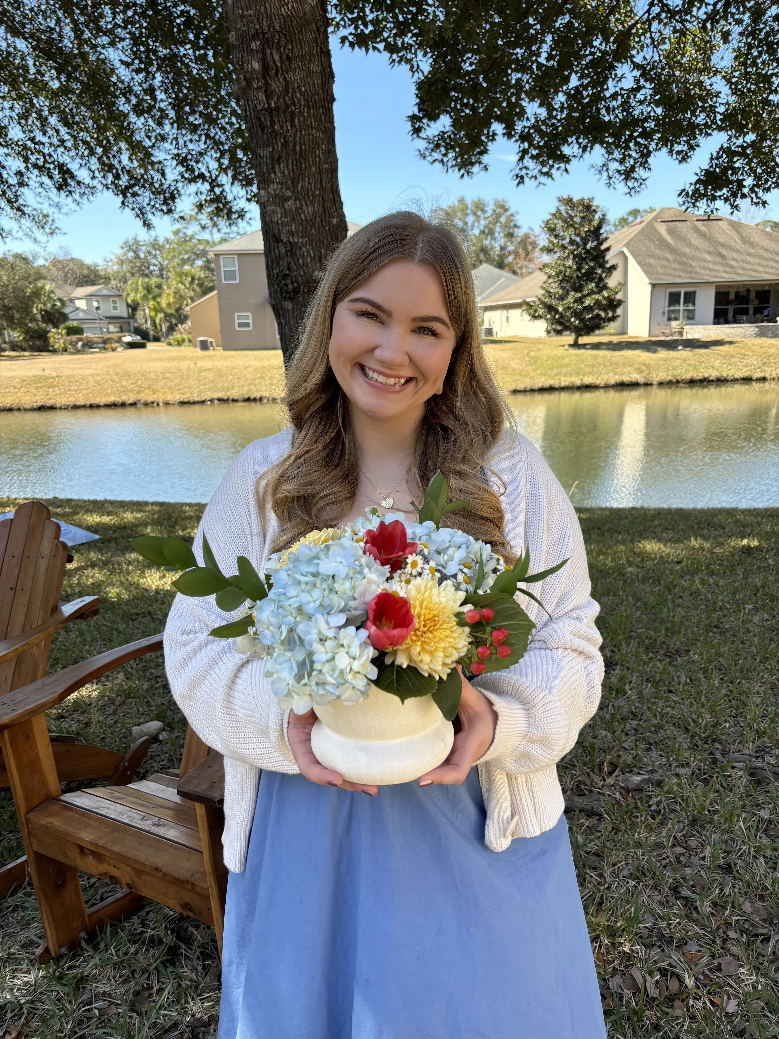 A young woman with long, wavy blonde hair smiling while holding a bouquet of colorful flowers in a white vase outdoors near a pond, with houses and trees in the background.