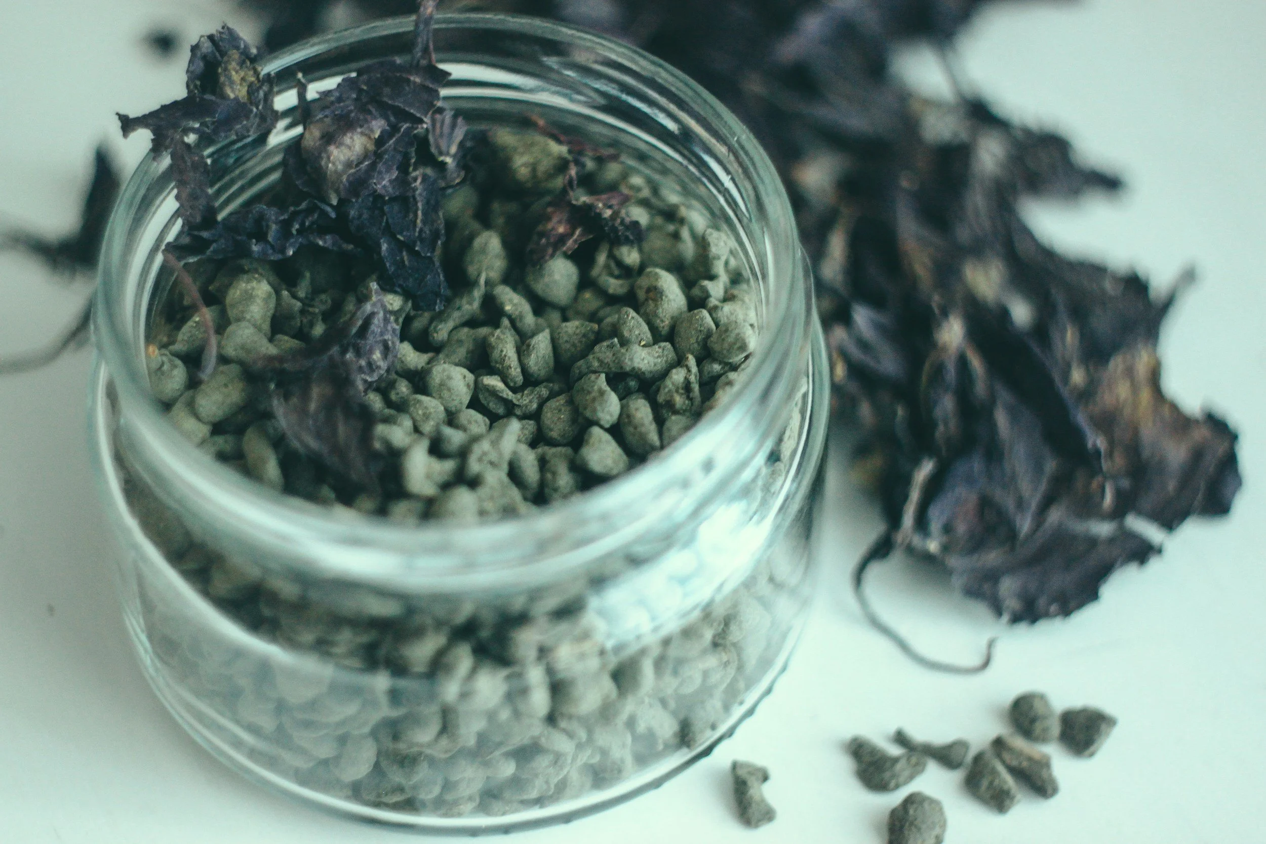 A glass jar filled with small gray pebbles and dried purple leaves next to it and some loose purple leaves on a white surface.