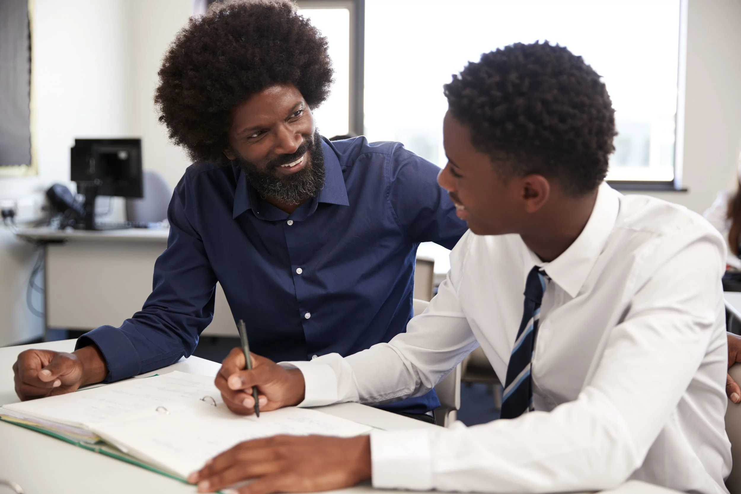 A teacher and a student are sitting at a desk in a classroom. The teacher is leaning over, talking to the student who is smiling and holding a pencil over an open notebook.