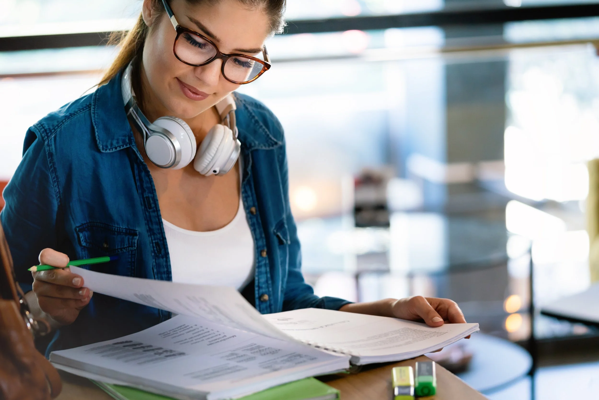A young woman with glasses and headphones around her neck studying documents at a table in a bright indoor setting.