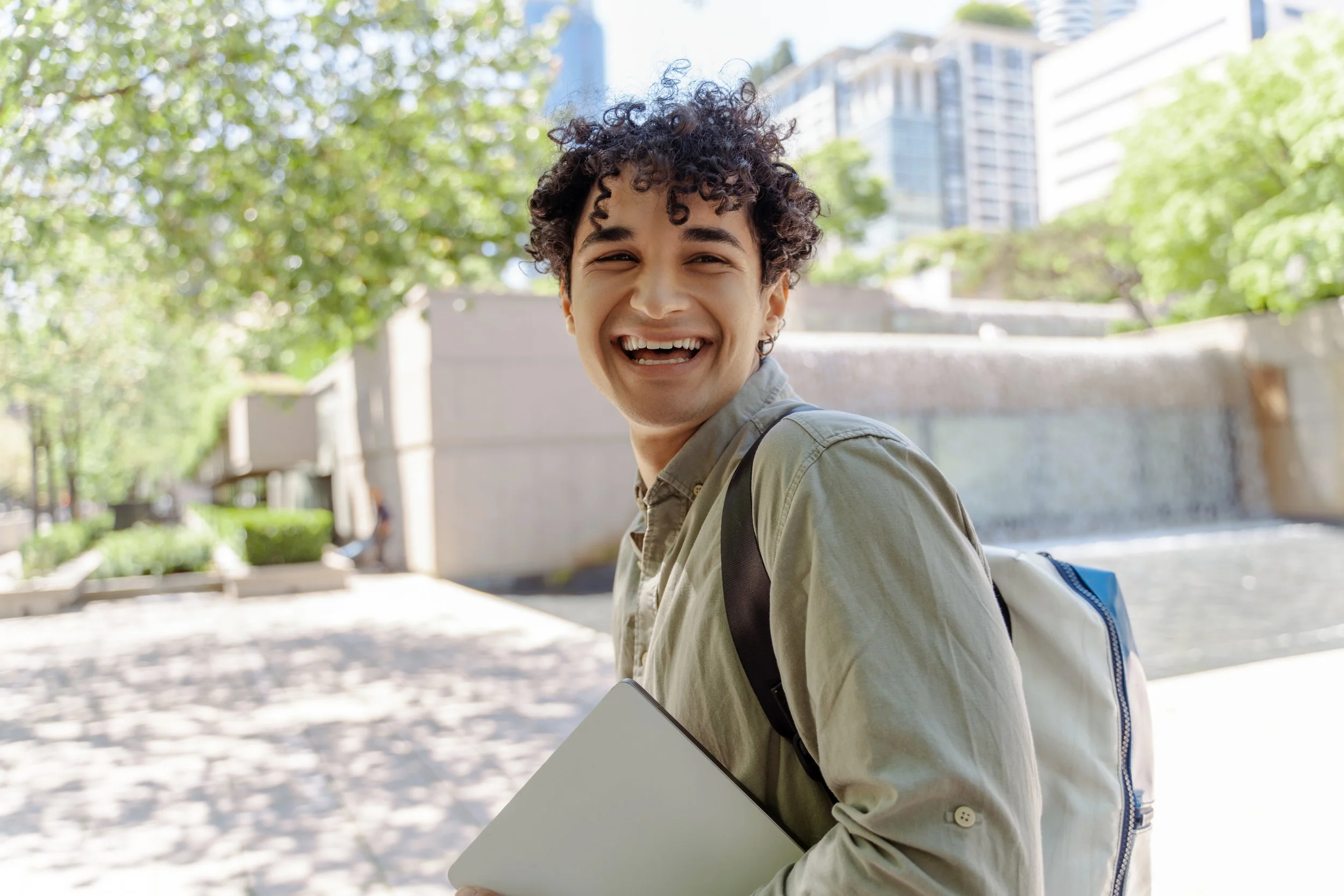 A young person smiling outdoors on a sunny day, carrying a backpack and holding a laptop in front of a water feature with greenery and skyscrapers in the background.