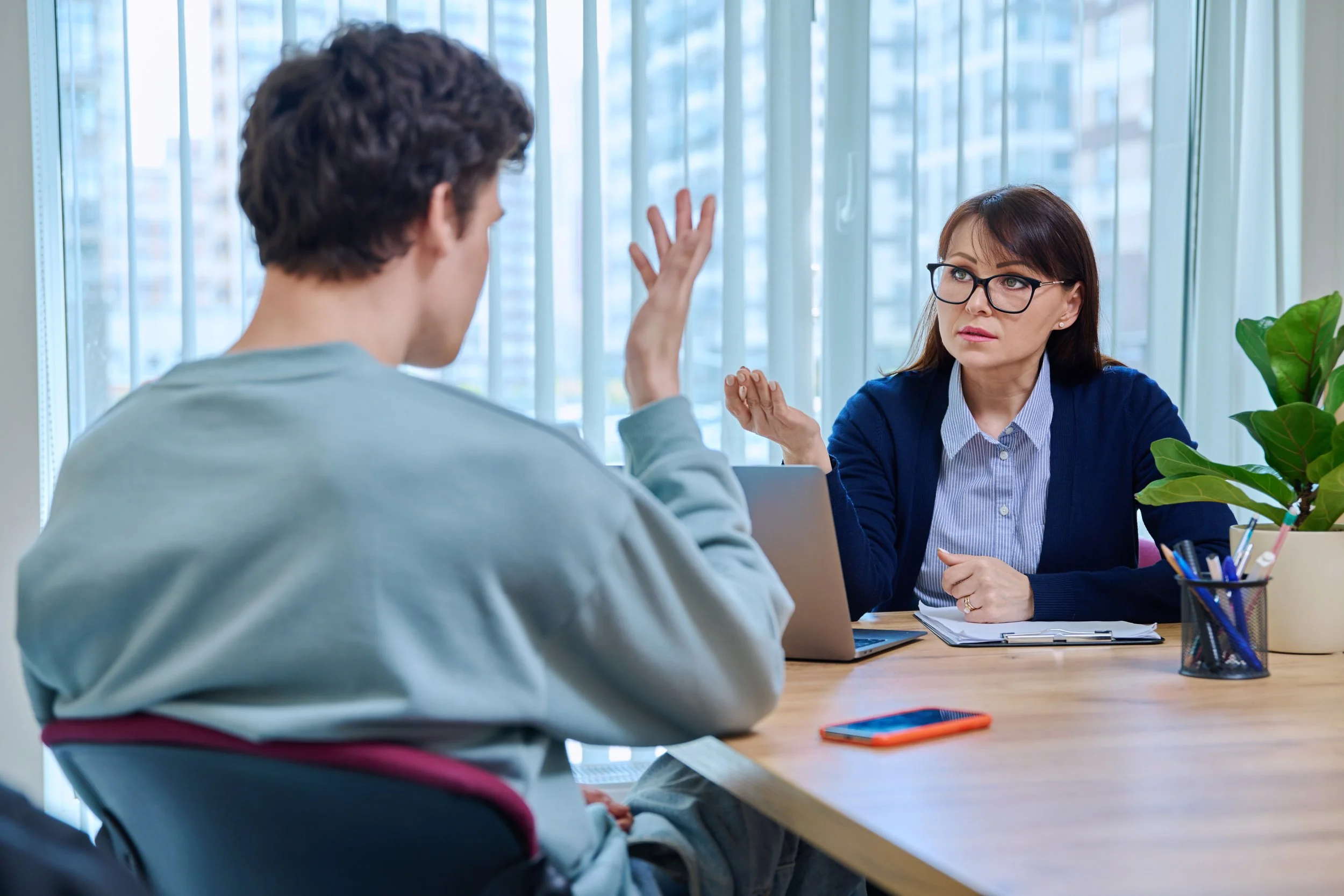 A woman in glasses and a blue cardigan sitting across from a young man with dark hair, discussing or explaining something during a meeting or consultation in an office with large windows and a city view.