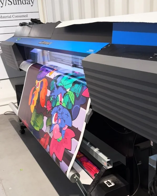 A large-format printer producing a colorful, cartoon-style print featuring various creatures and shapes on a sheet of paper.