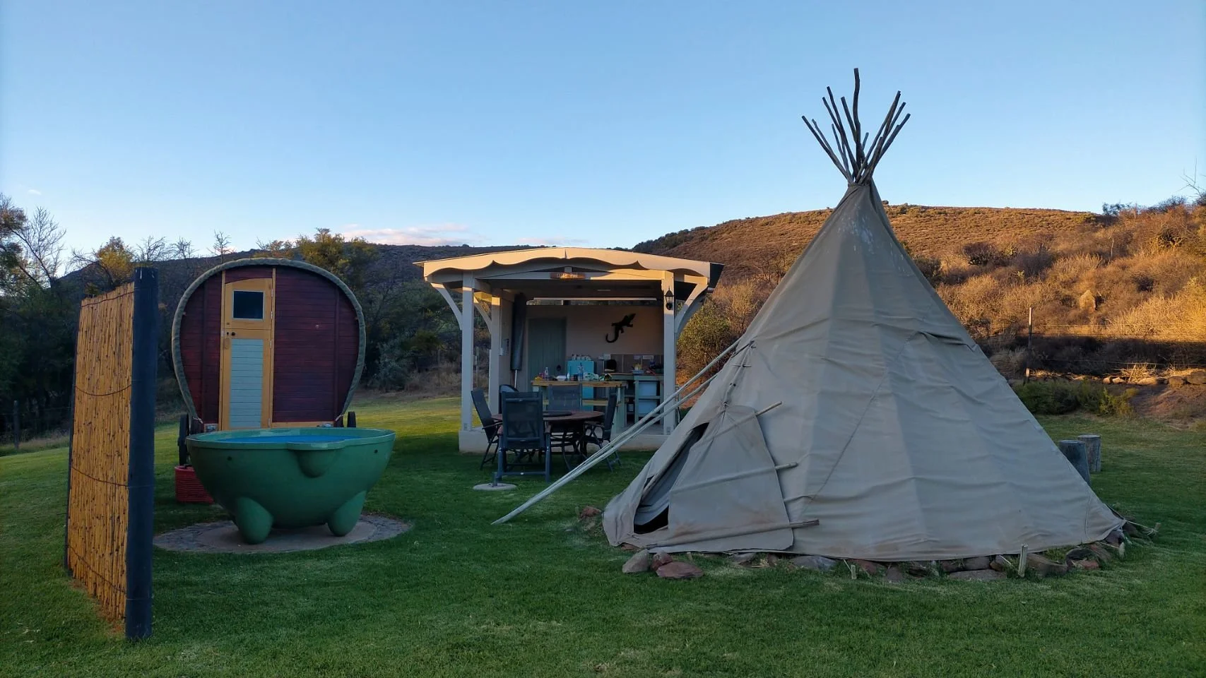 A teepee en toile beige avec une structure en bois au sommet, une petite cabine en bois rouge foncé avec une porte jaune, une baignoire verte en plastique avec quatre pieds, un espace extérieur avec un mobilier de cuisine et une table de repas sous un abri avec un mur décoré d’un lézard noir, dans un paysage naturel avec des collines et un ciel clair.