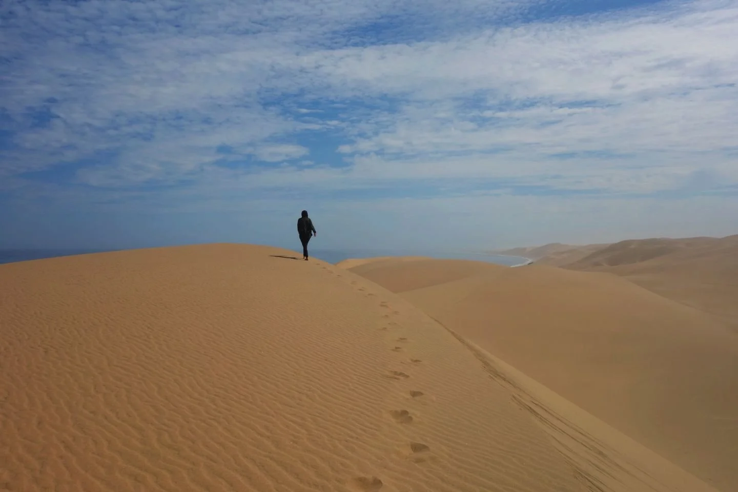 Une personne marche sur une dune de sable dans un désert, laissant des traces derrière elle, avec un ciel partiellement nuageux au-dessus.