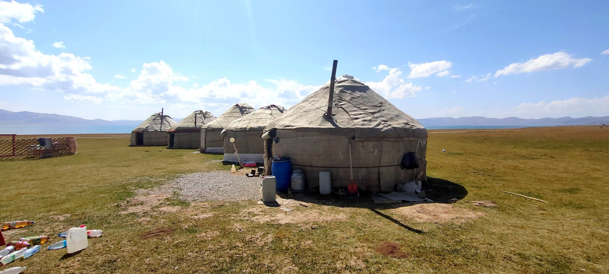 Un groupe de yourtes en toile beige installé sur un paysage de prairie avec vue sur un lac et des montagnes au loin, sous un ciel bleu avec quelques nuages.