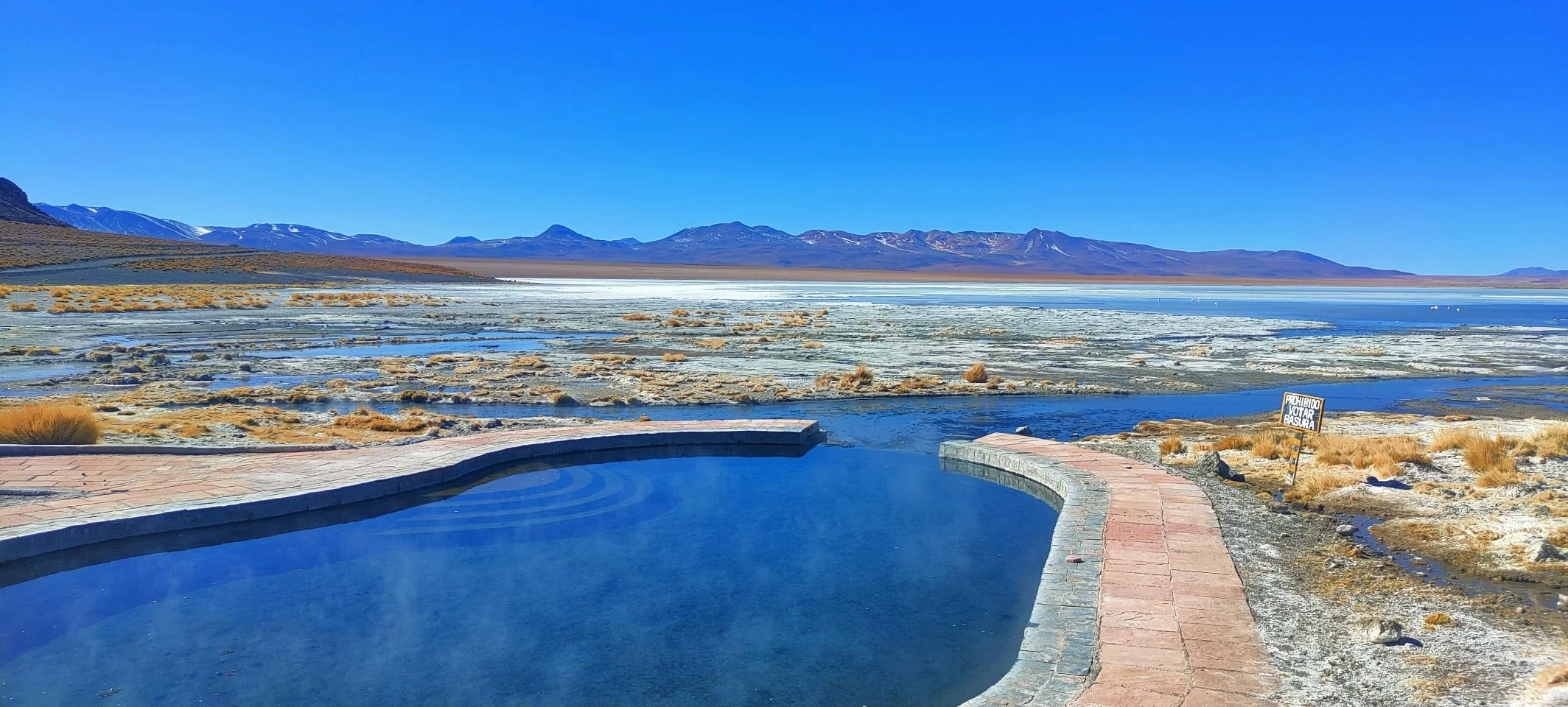Vue d'une source chaude dans un paysage désertique avec des montagnes en arrière-plan, ciel bleu clair et un panneau qui indique "Prohibido votar basura".