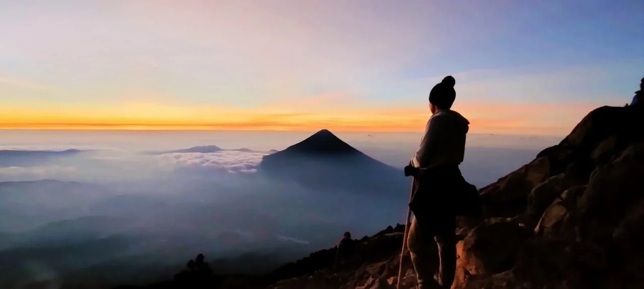 Une personne en vêtements chauds et chapeau se tient sur une pente rocheuse au coucher du soleil, avec un volcan en arrière-plan et un ciel coloré.