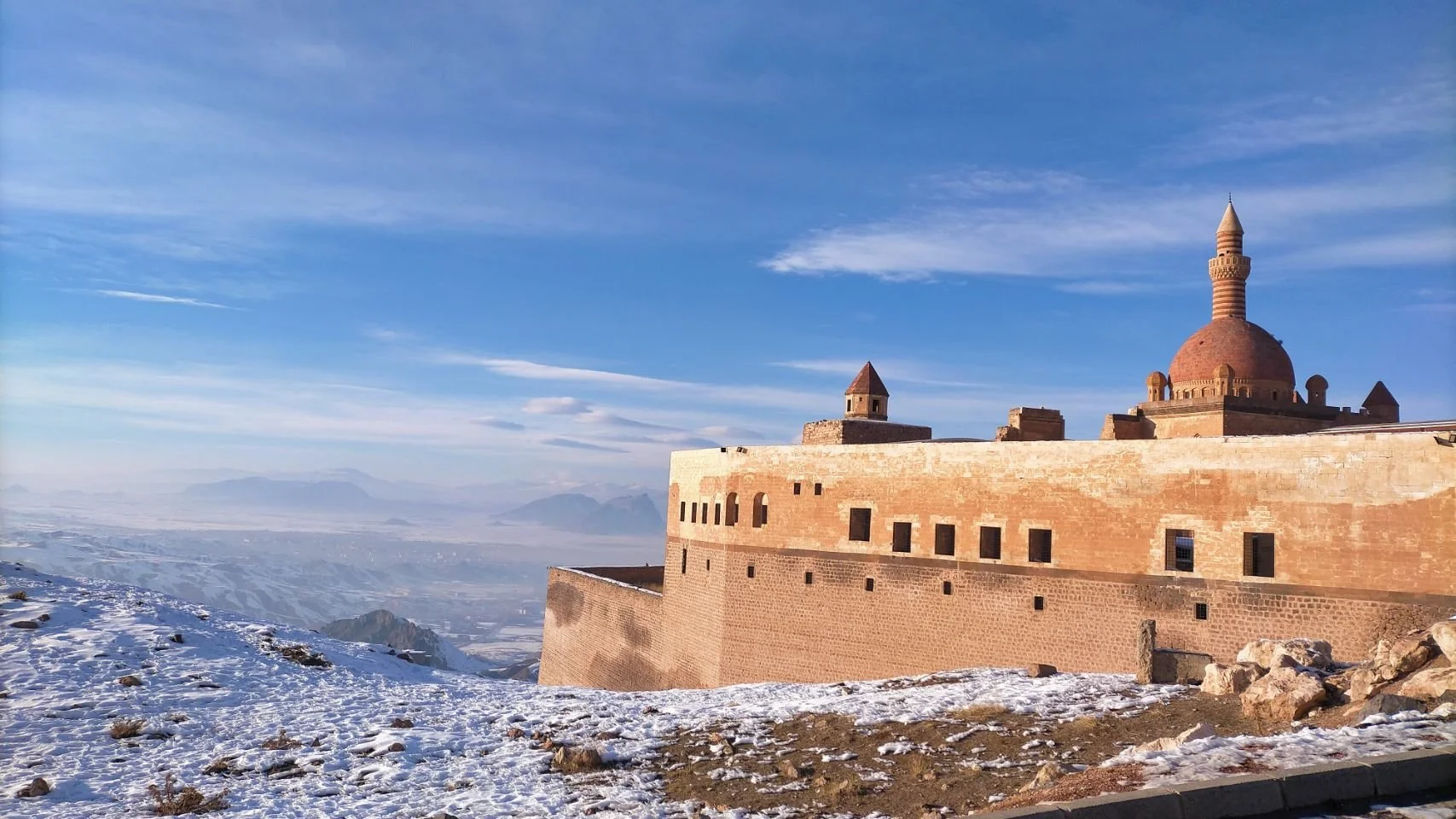 Vue d'une forteresse en pierre en altitude avec un ciel bleu et quelques nuages, neige sur le sol en premier plan, montagnes à l'horizon.