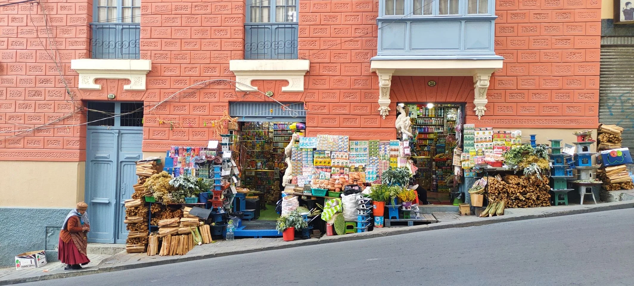 Magasin de rue coloré avec des produits en vente à l'extérieur, notamment des branches de bois, des fleurs, des jouets, des boîtes, et des plantes, avec une femme âgée portant un foulard et un manteau en train de marcher sur le trottoir.