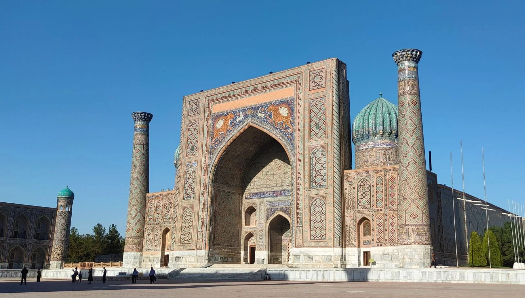 Une grande porte décorée avec des motifs islamiques et deux minarets, flanquée d'un bâtiment en pierre, sous un ciel bleu clair.