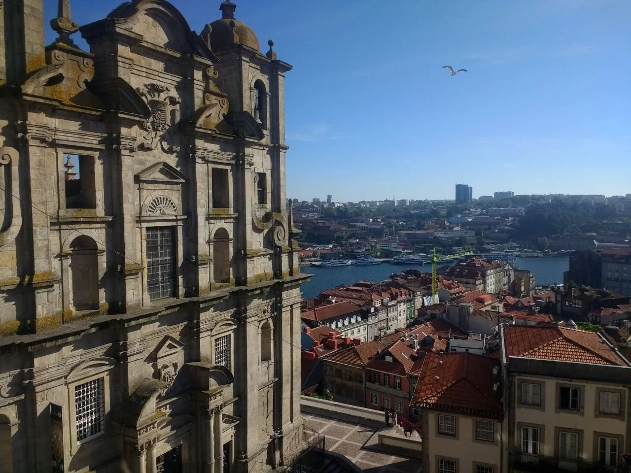 Vue aérienne d'un bâtiment historique en pierre, avec une ville et un fleuve en arrière-plan, sous un ciel clair avec un oiseau en vol.
