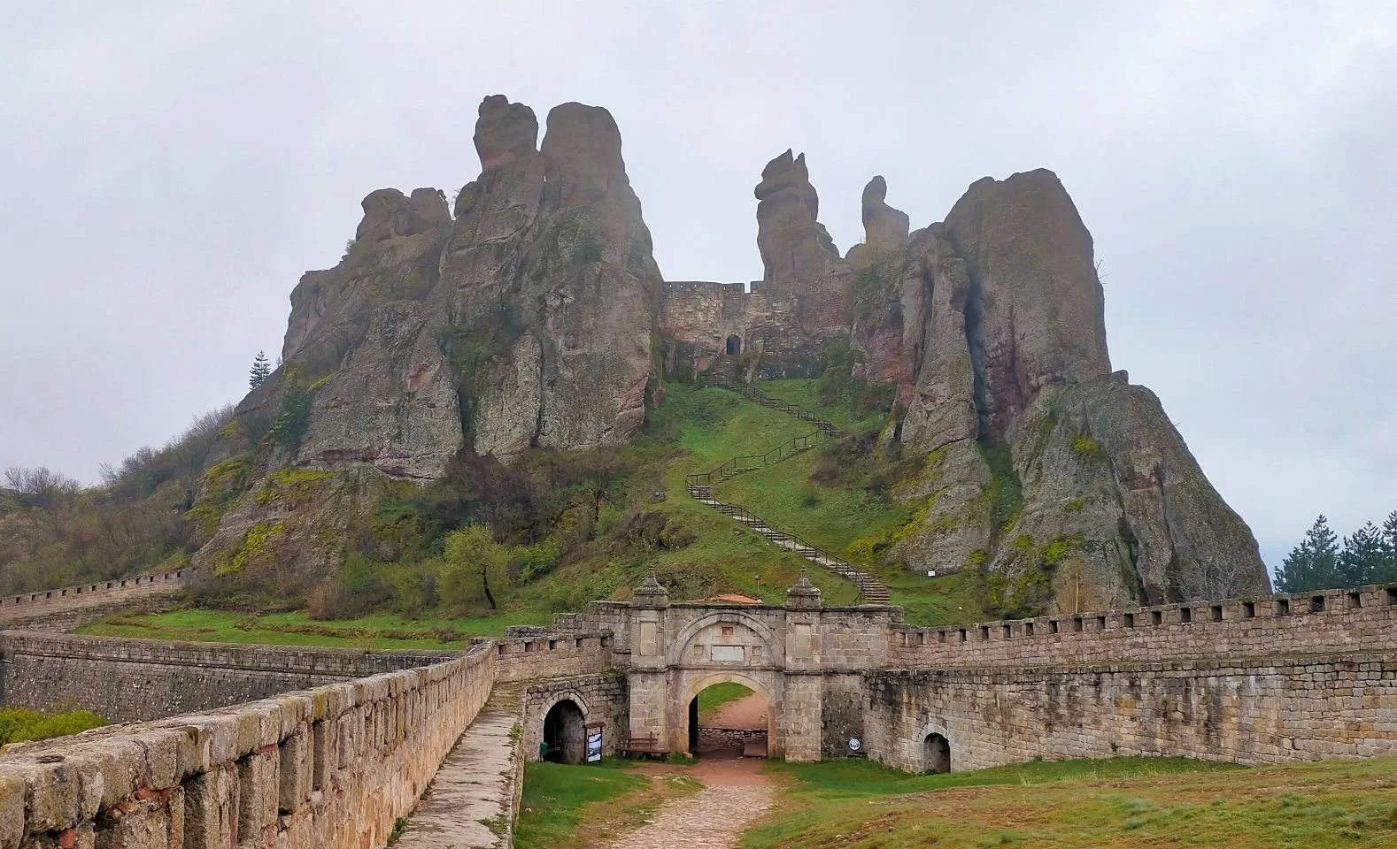 Ancienne forteresse en pierre construite au pied d'une formation rocheuse montagneuse accidentée, avec une entrée principale et un chemin en montée menant au sommet.