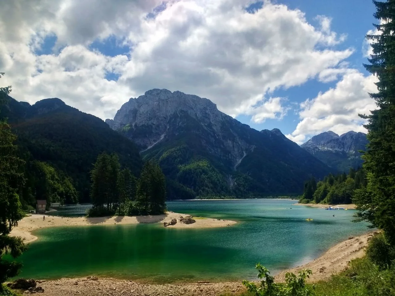 Lac entouré de montagnes avec un ciel partiellement nuageux, des arbres vert foncé et une petite plage de sable.