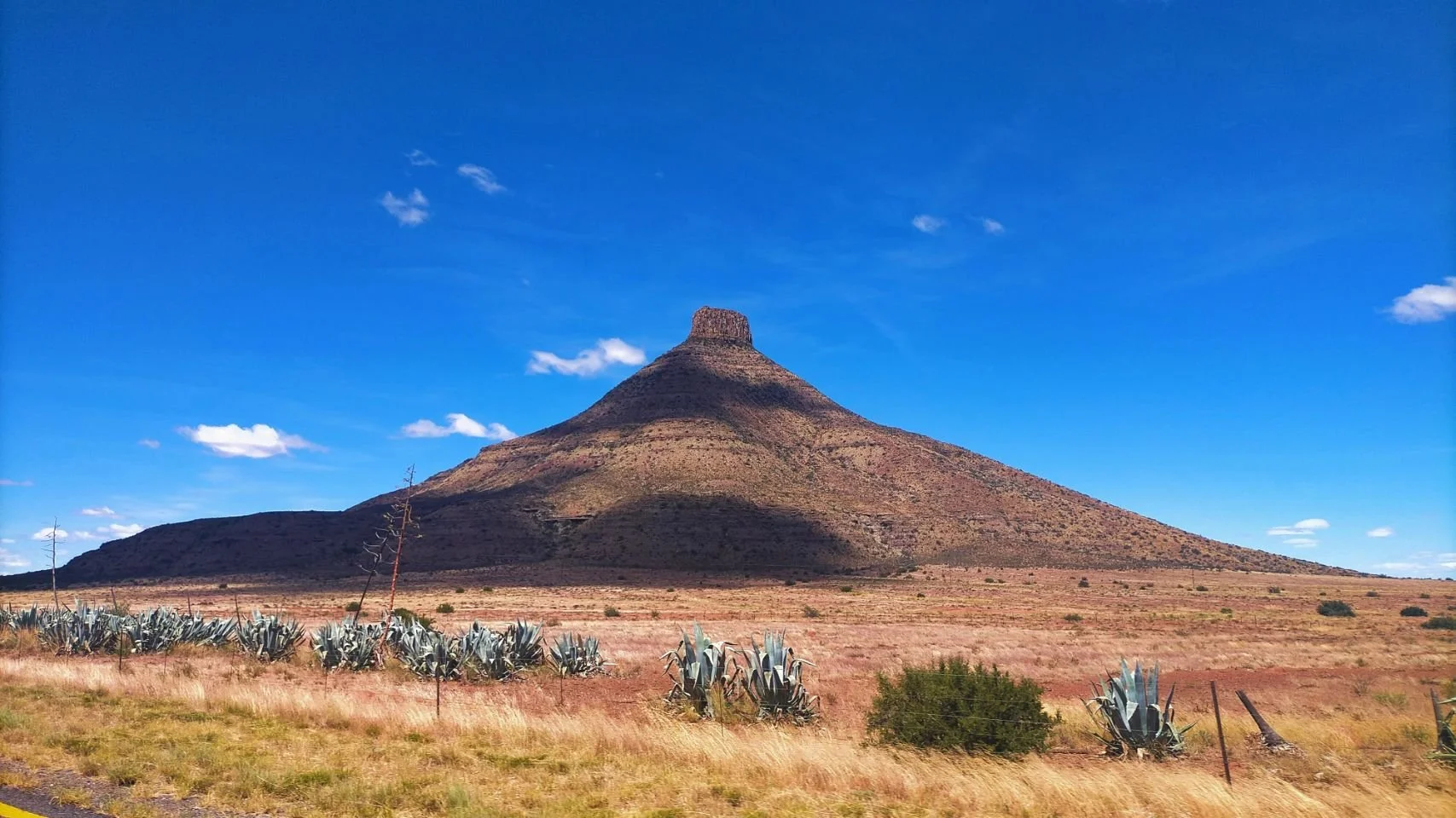 Montagne en forme de pyramide avec sommet plat dans un paysage désertique aride, ciel bleu avec quelques nuages, plantes succulentes et buissons au premier plan.