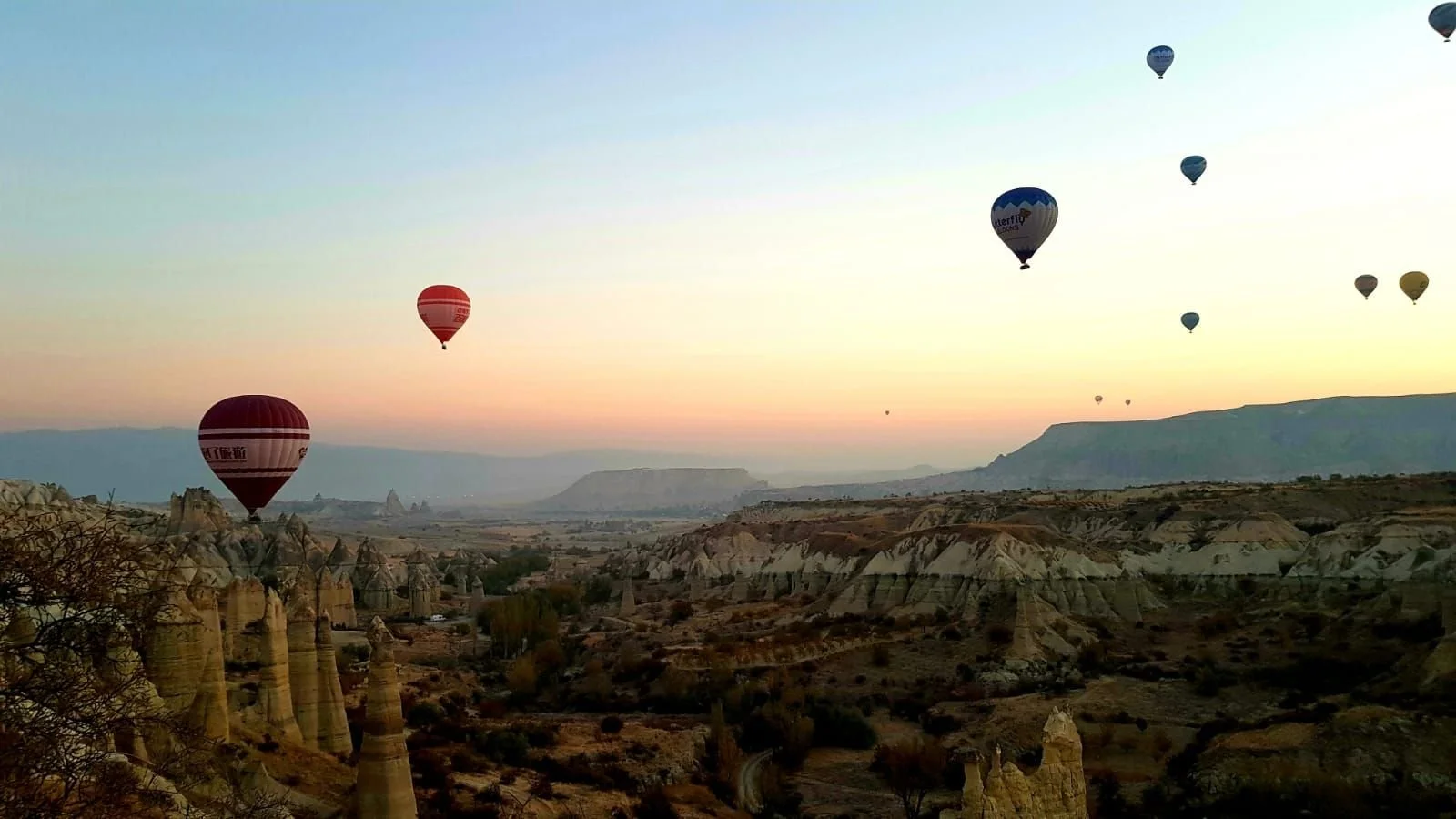Plusieurs montgolfières colorées flottent au-dessus d'un paysage rocheux au lever du soleil, avec des formations géologiques uniques.
