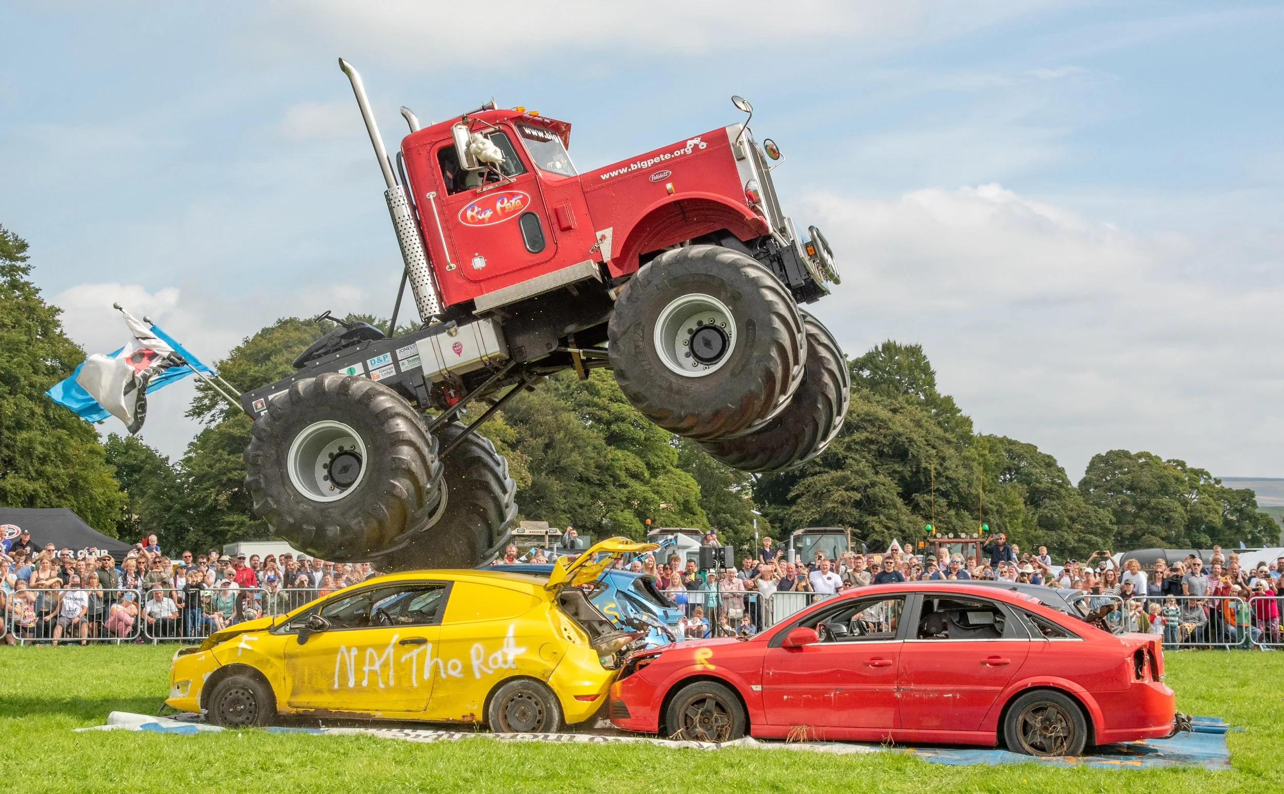 Large red monster truck jumping over two wrecked cars, with spectators watching in the background.