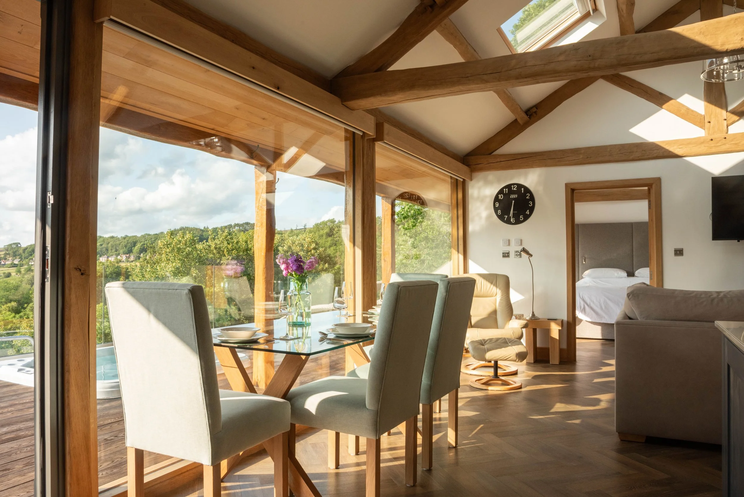 A bright, sunny living and dining area with large glass sliding doors revealing a view of greenery and a blue sky. The room features wooden beams and flooring, a glass dining table with four chairs, and a cozy seating area with armchairs and a wall clock.