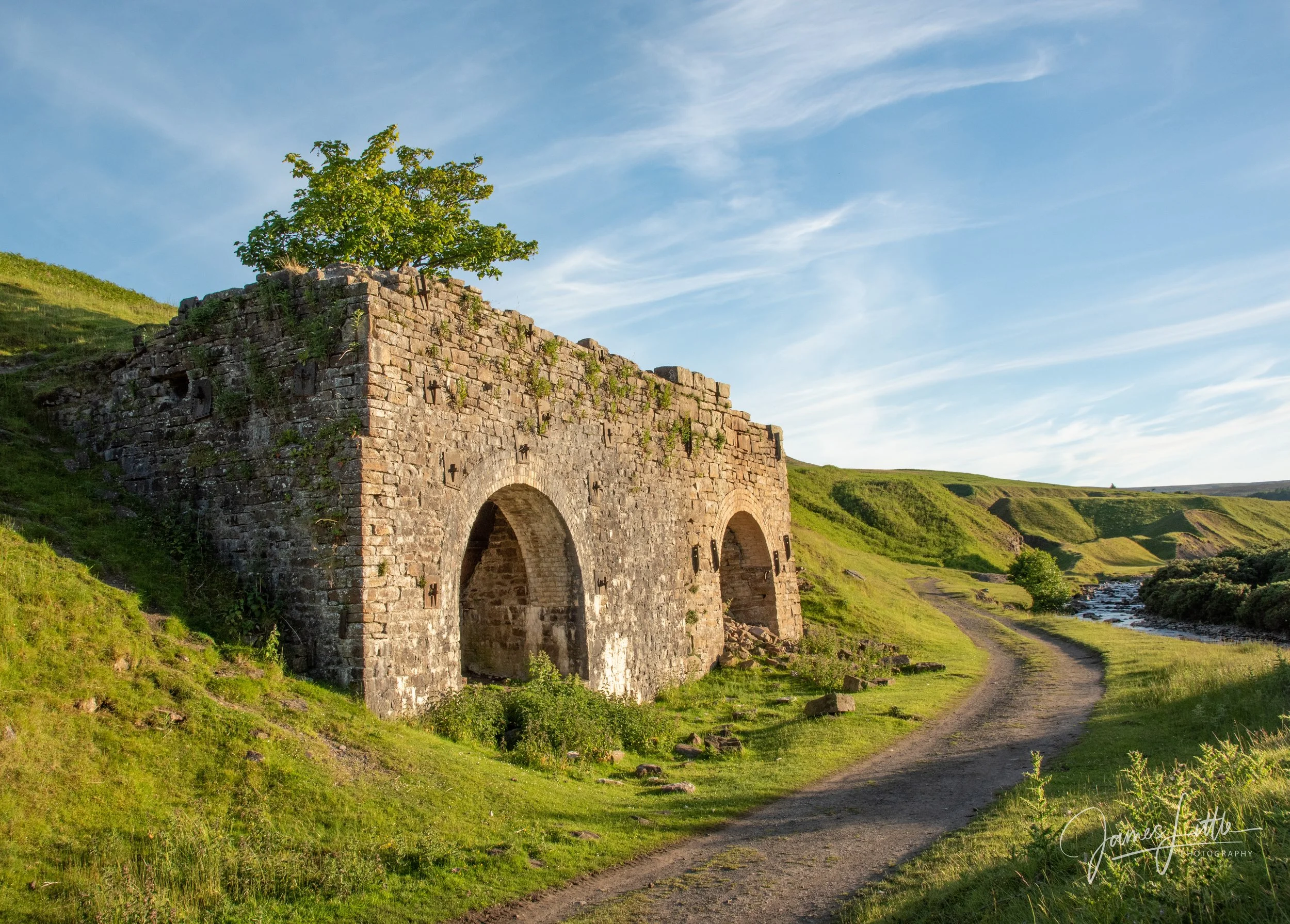 A stone arch bridge with a tree on top, set in a grassy, hilly landscape under a blue sky with wispy clouds, and a dirt path leading past the bridge along a river. An old lime kiln at Bollihope near Frosterley. 