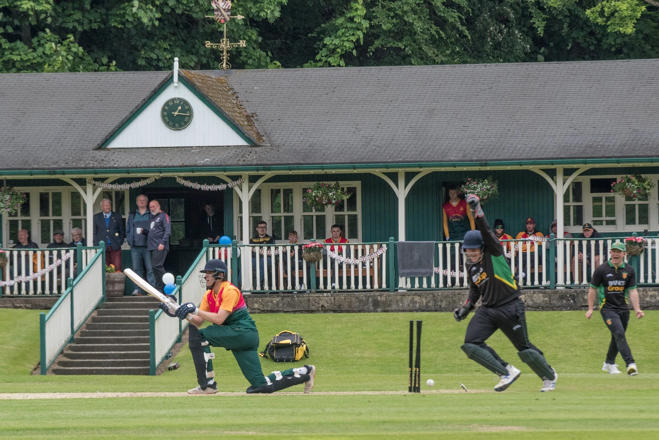 Cricket players on the field, with one player kneeling, another advancing, and a third preparing to bat, in front of a pavilion with spectators and officials sitting and standing.