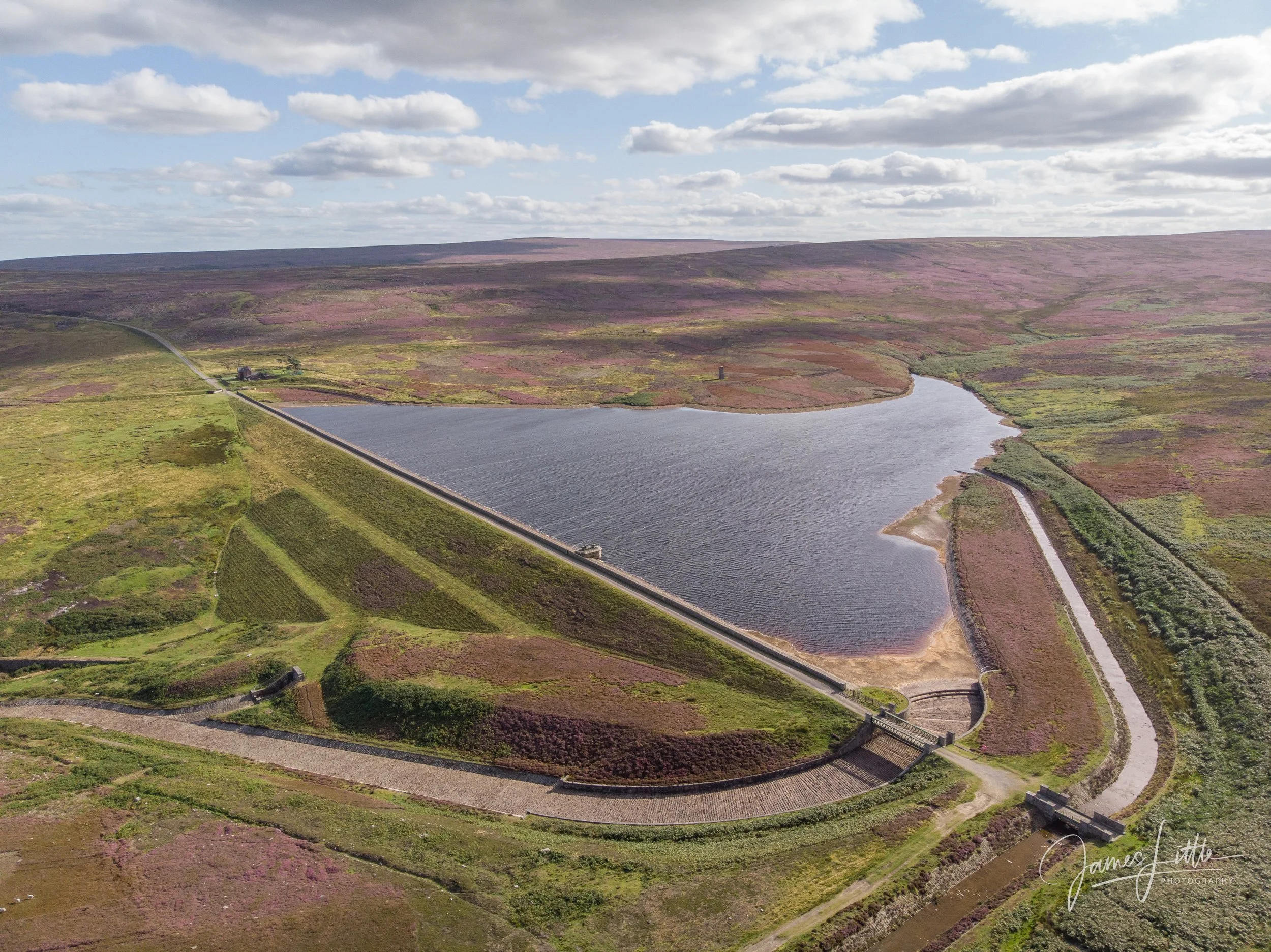 Aerial view of a large reservoir with dam walls on both ends, surrounded by green and purple heathland fields, with a sky full of clouds. This is Hisehope reservoir near the waskerly way in County Durham. 