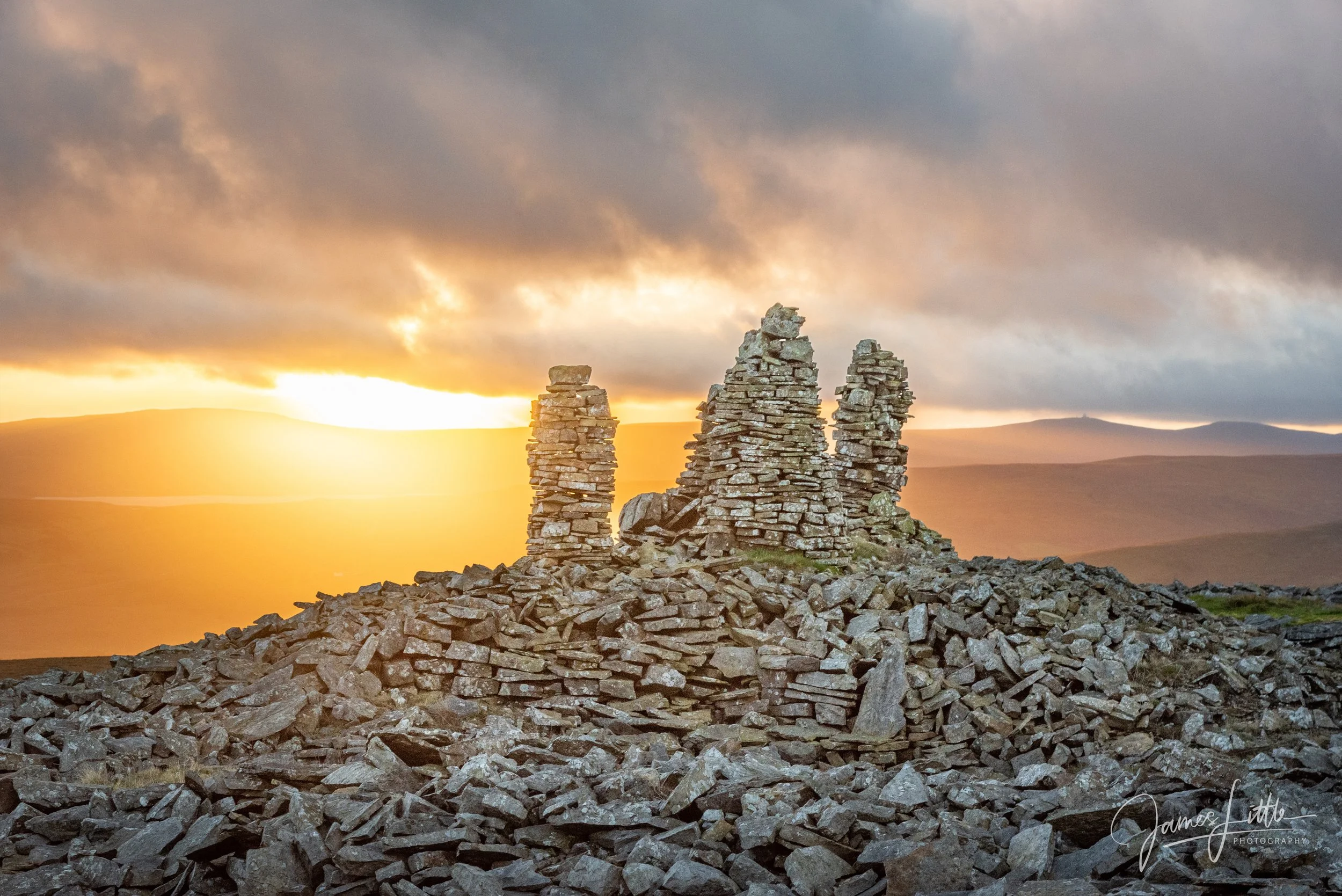 Sunset over rolling hills with several stacked stone cairns on rocky terrain in the foreground.