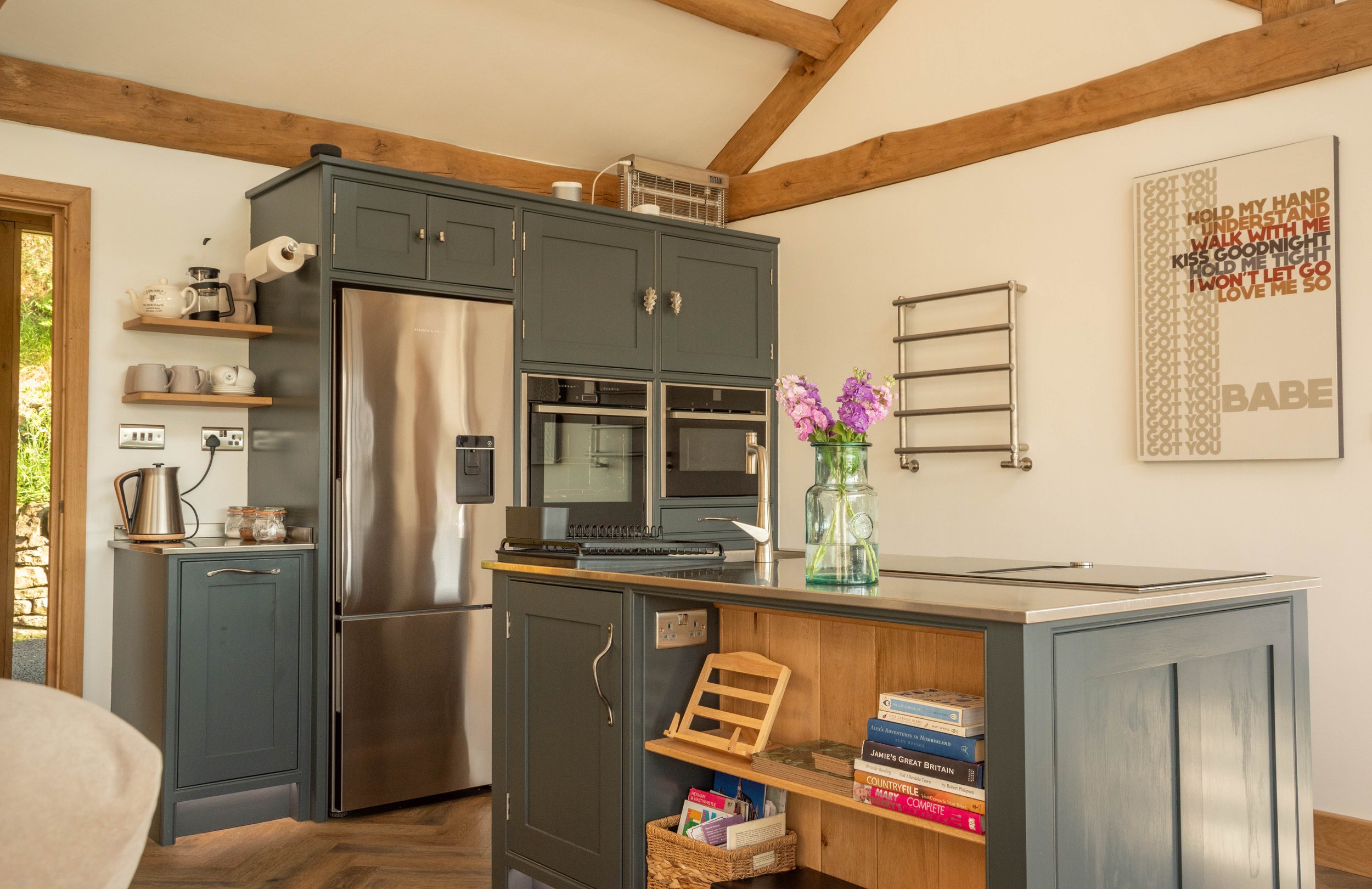 A cozy kitchen with blue cabinetry, a stainless steel refrigerator, an electric kettle, open shelves with cups and jars, a countertop with a vase of purple flowers, and wall art with song lyrics.