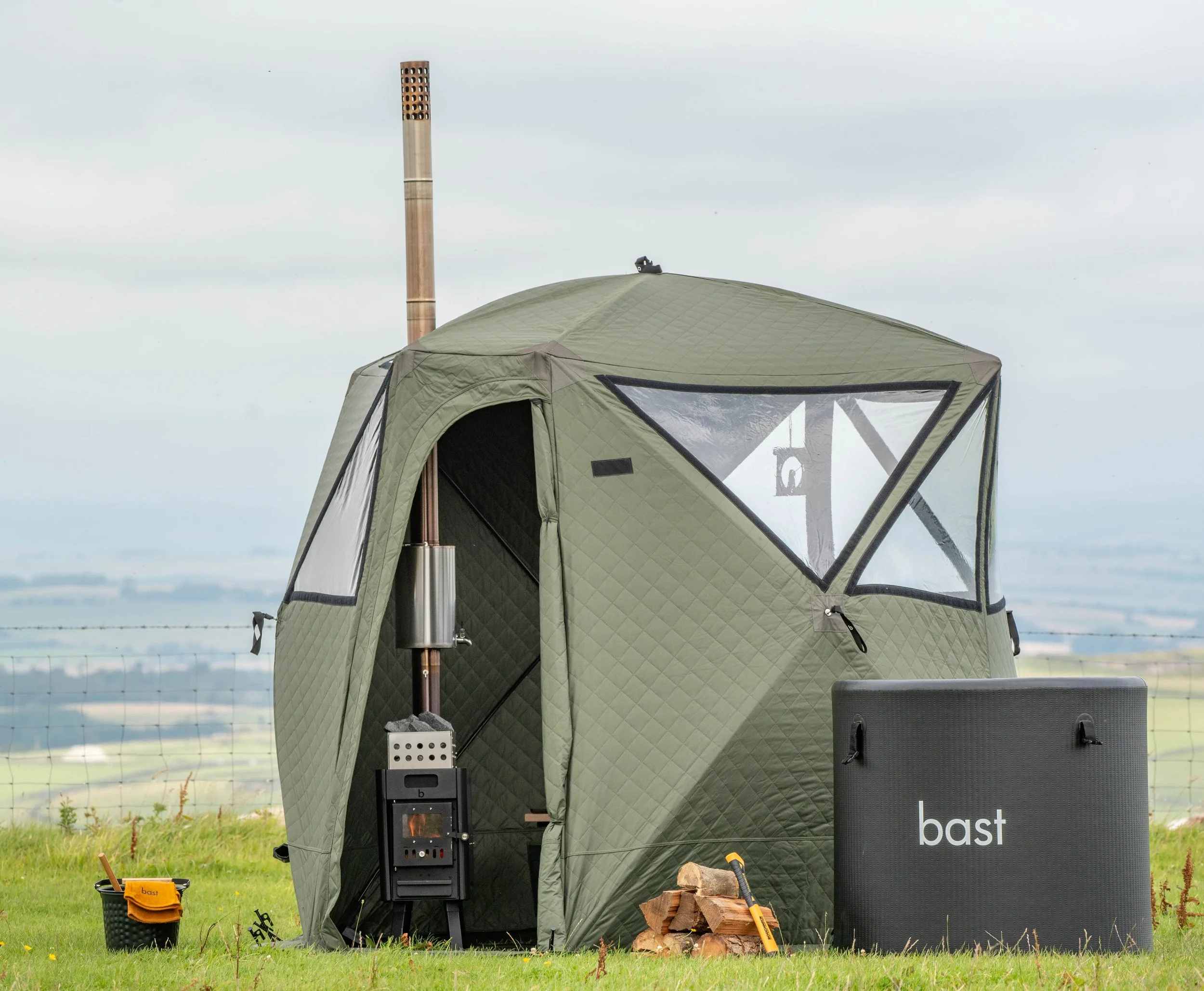 Green insulated outdoor tent set up on grassy field with a chimney, wood logs, and outdoor stove, surrounded by outdoor gear and equipment, with a barbed wire fence and landscape in the background.