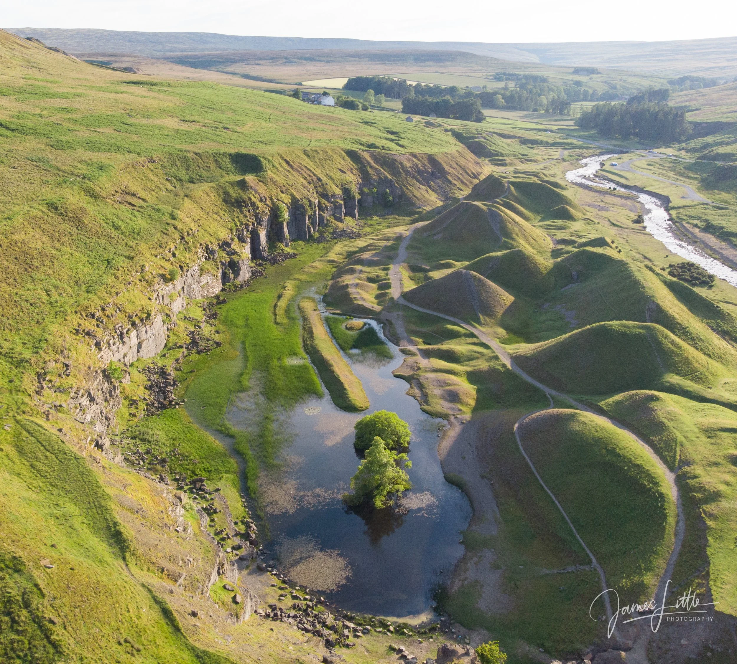 A green, hilly landscape with a winding river and small patches of trees, seen from an aerial perspective. Bollihope quarry during the summer months. 