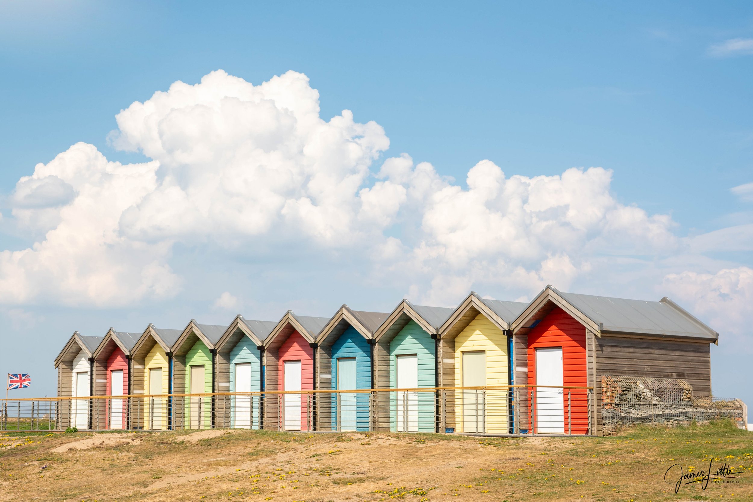 Row of colorful beach huts on a grassy hill with a cloudy sky and a small British flag in the background.