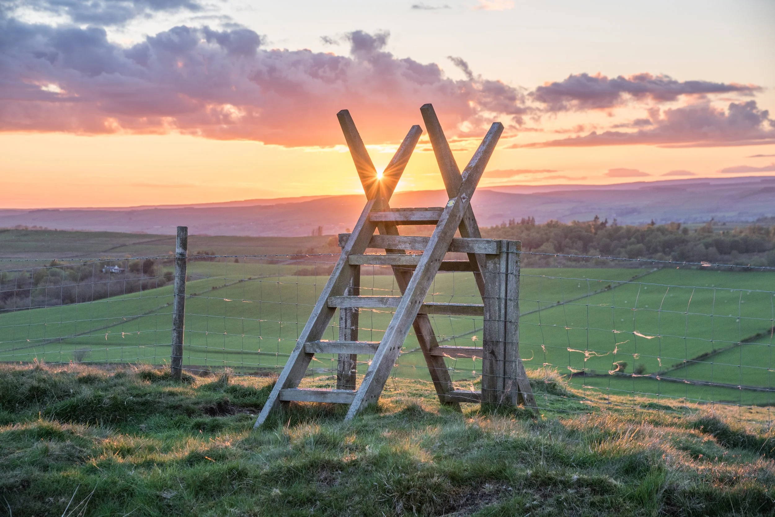 Wooden ladder leaning on a fence post in a rural landscape at sunset, with green fields and hills in the background.