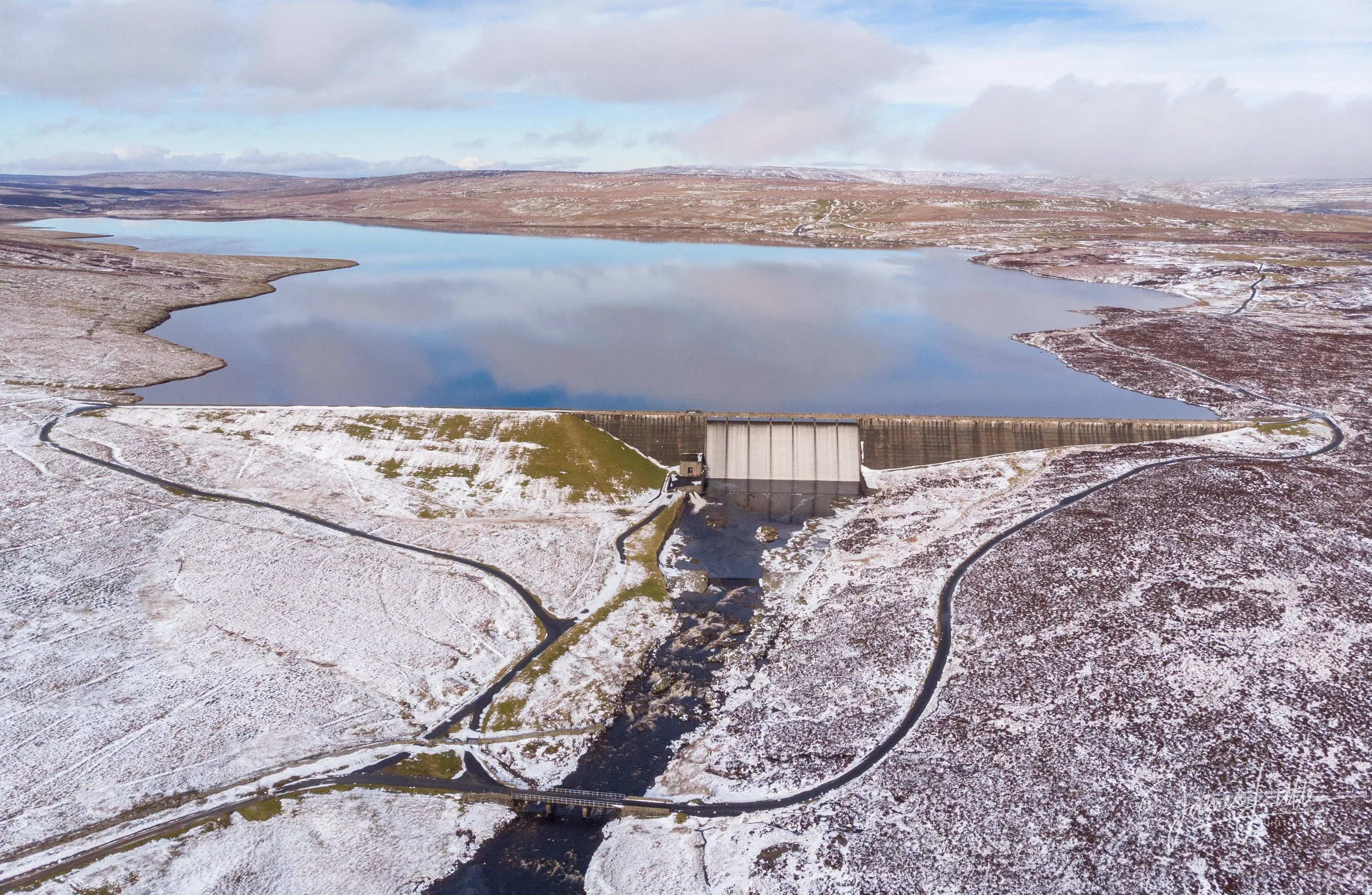 A dam on a partially frozen lake in a snowy landscape, with winding roads and hilly terrain in the background. Cow Green Reservoir overflowing following lots of rain. 