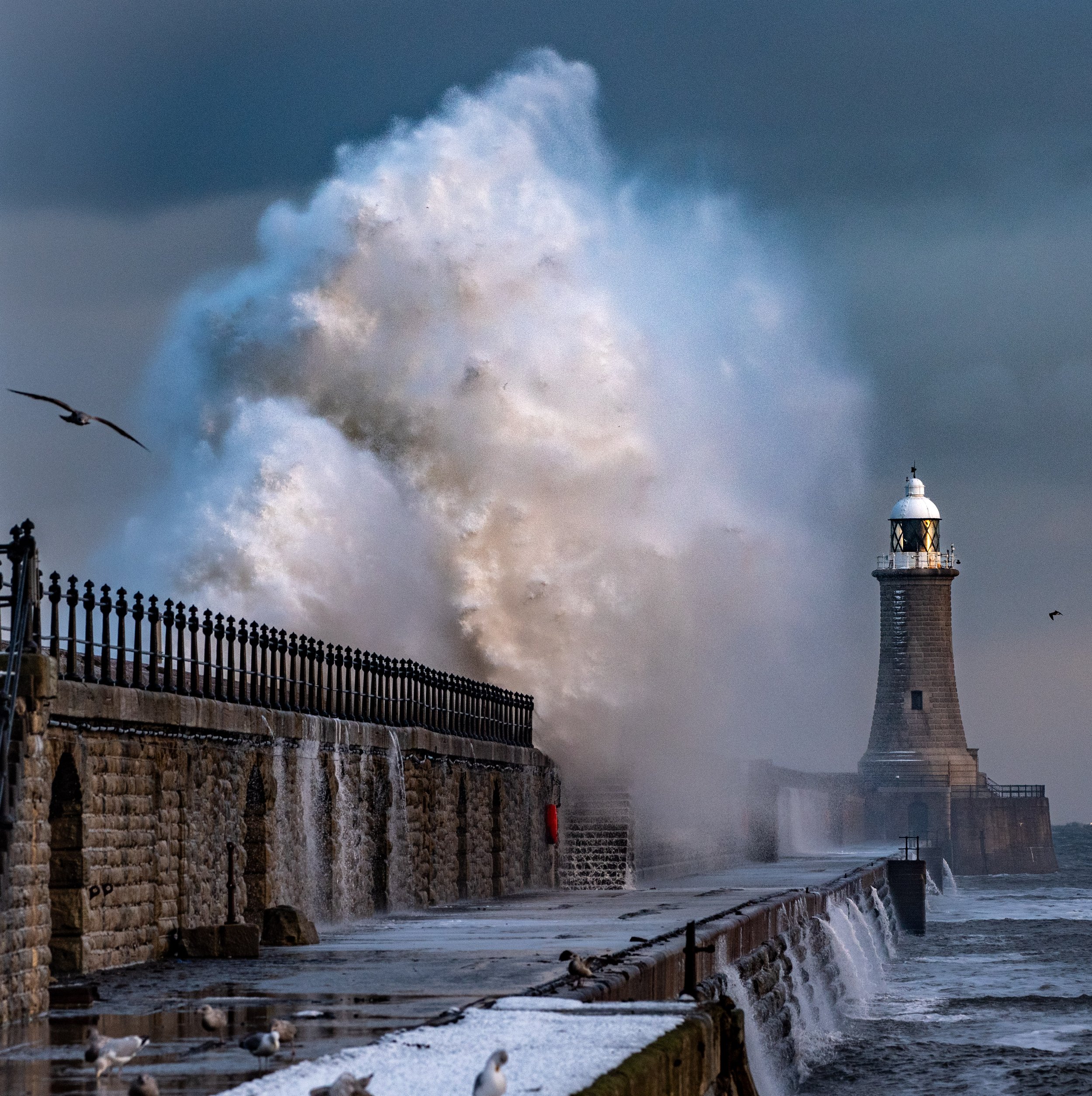 Ocean waves crashing against a lighthouse and seawall, with seagulls flying overhead on a cloudy day.