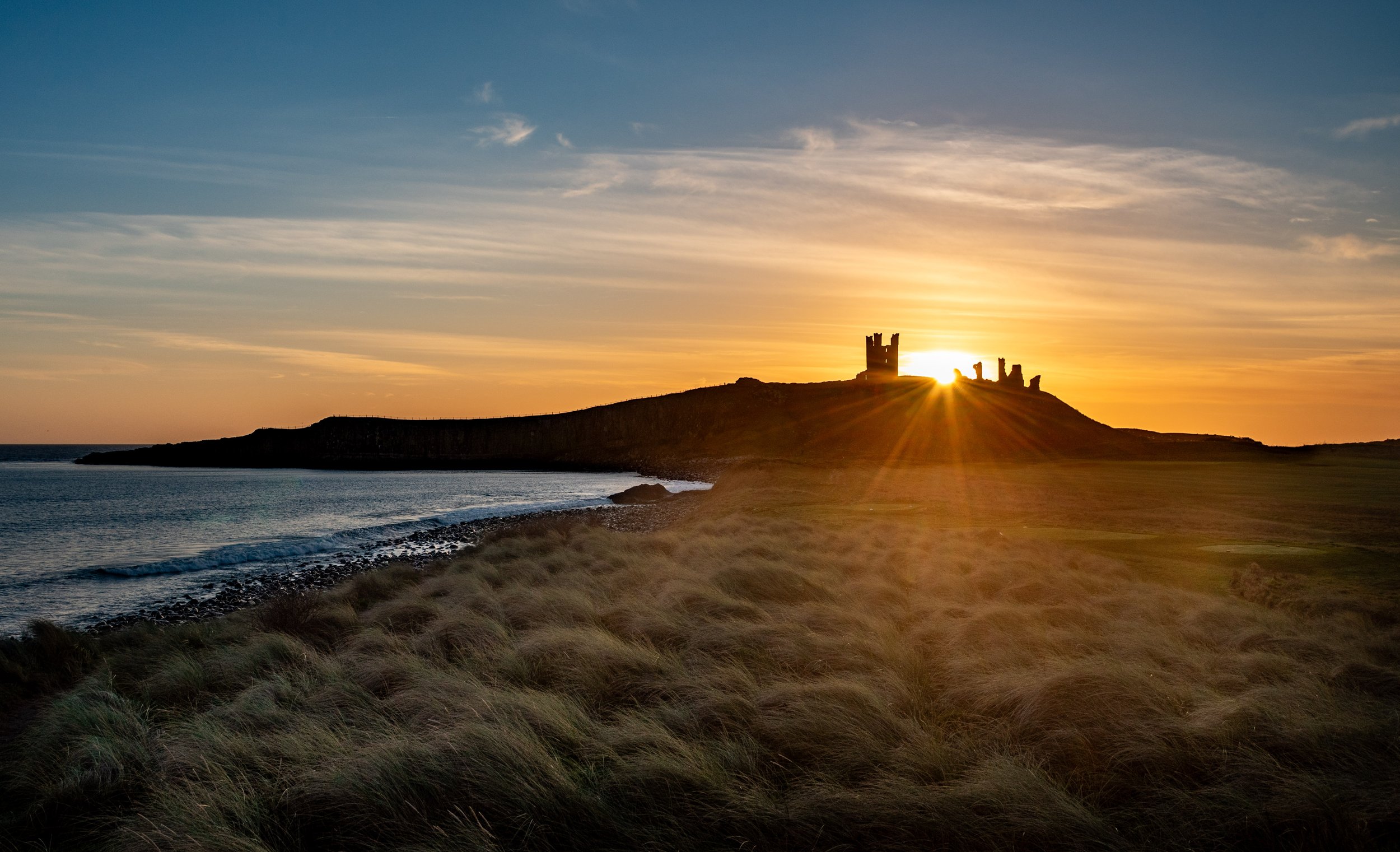 Sunset over a coastal landscape with grassy dunes and a castle silhouette on a hill.