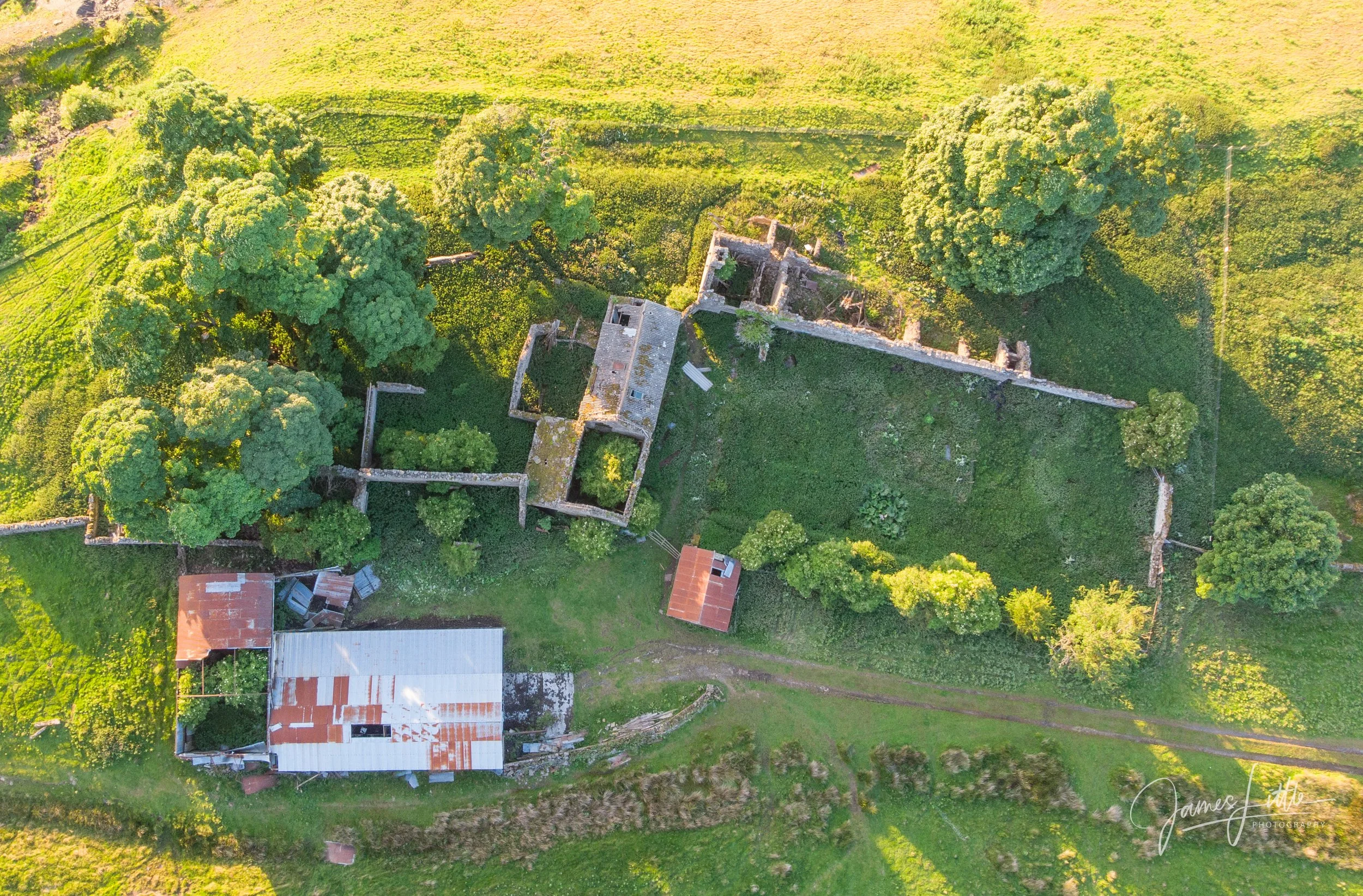 An aerial view of an abandoned house surrounded by green trees and grass, with a white metal-roofed building nearby. This old farm is just outside of Stanhope. 