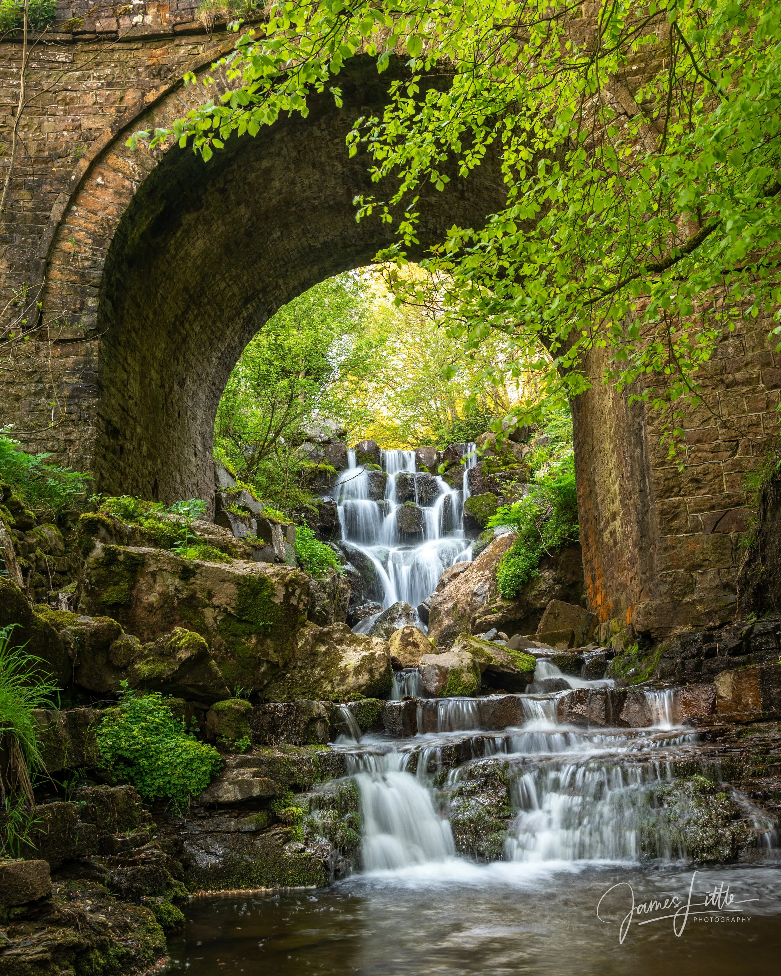 A lush green forest with a flowing waterfall beneath a stone arch bridge. The location is near Garrigill. 