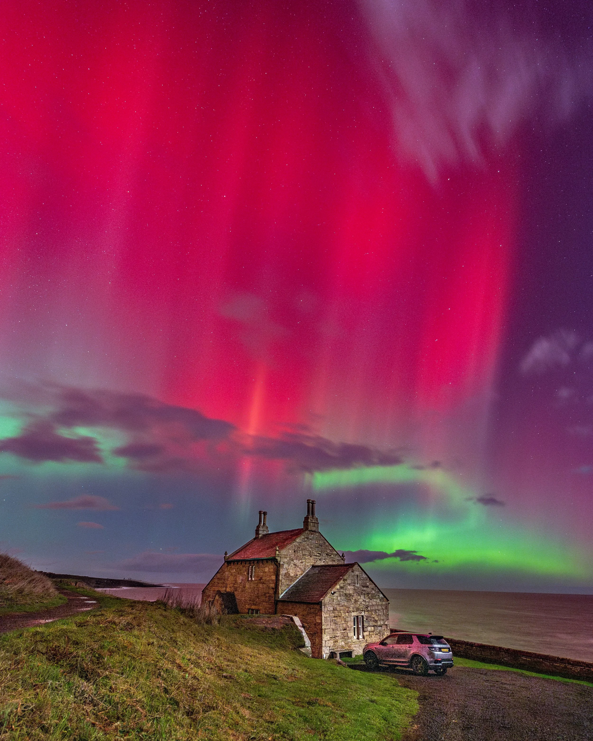 A house near the coastline under a colorful display of the northern lights with pink, green, and purple hues in the sky.