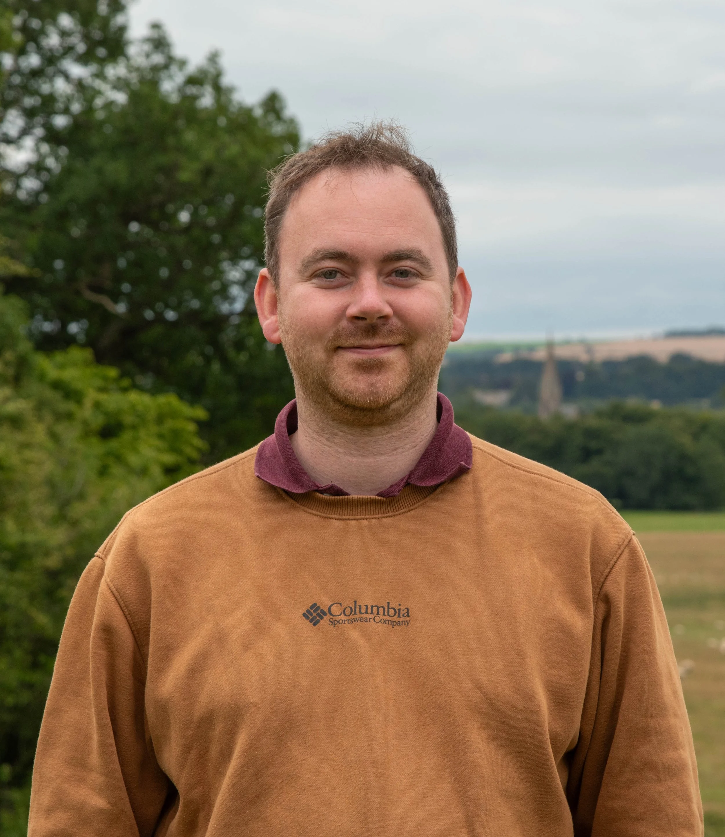 A man outdoors standing in front of a landscape with trees and a cloudy sky, wearing a brown Columbia Sportswear sweatshirt and smiling.