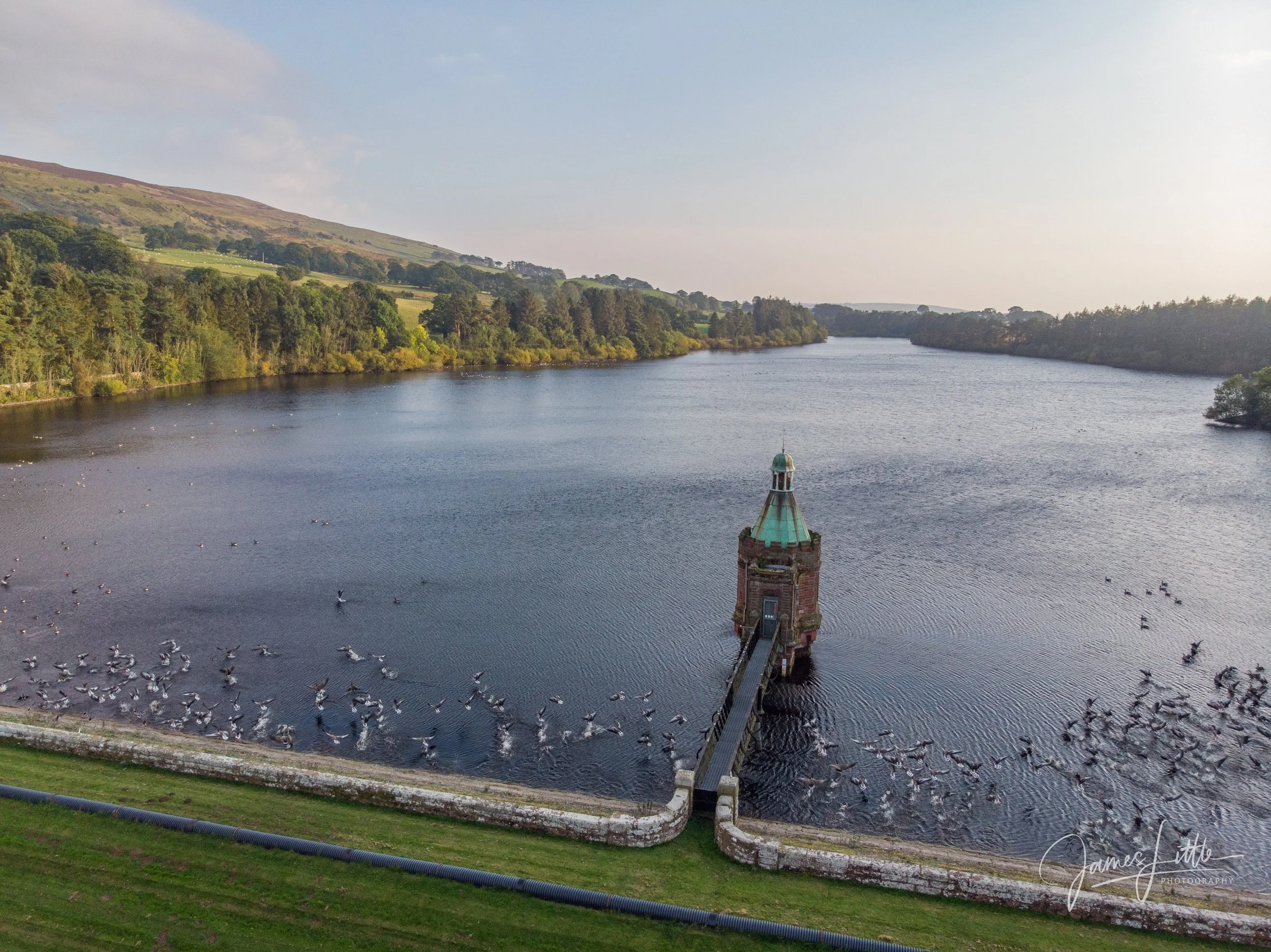 A large body of water with a small tower structure connected by a bridge in the foreground, surrounded by trees and hills in the background. Castle Carrock is the nearest town. 