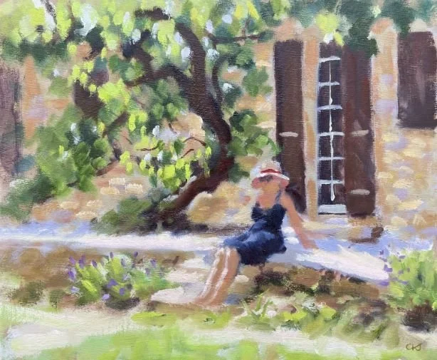 A woman in a navy dress and wide-brimmed hat relaxing on a wooden bench in a garden, with a tree and a brick house with windows nearby.