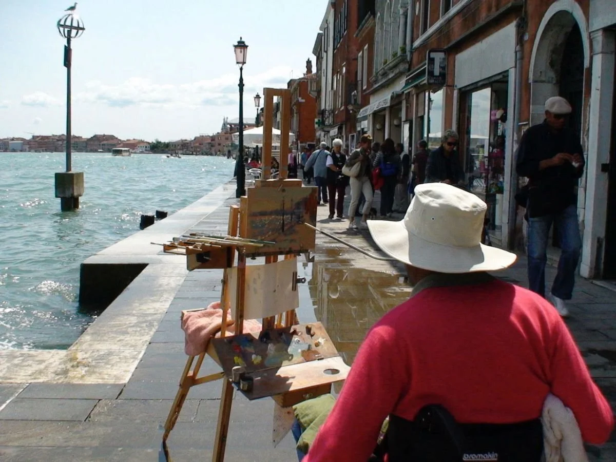 An artist painting at an outdoor waterway with people walking along a city street in the background.