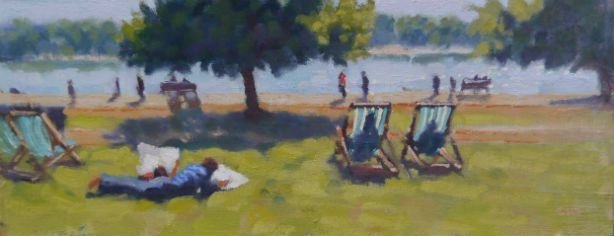 A person lying on the grass next to five empty lounge chairs near a lake, with trees and people walking along the shore in the background.