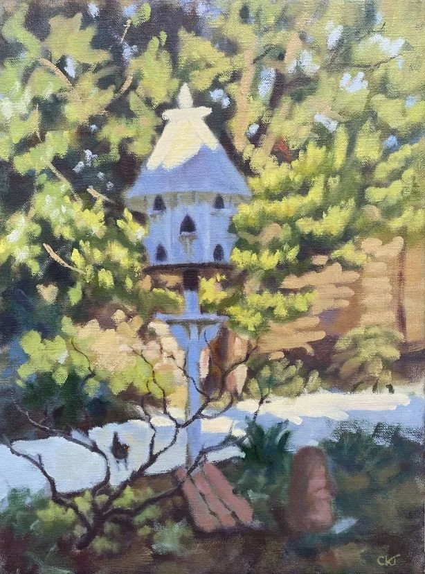 A painting of a birdhouse in a tree, with a pond and rocks below and a wooden bench nearby.