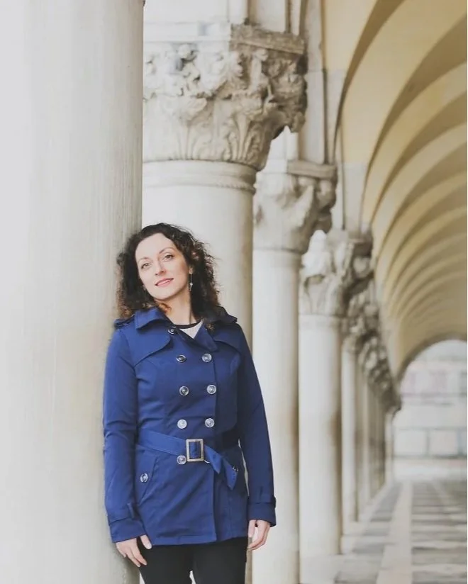 Une femme avec des cheveux bruns bouclés portant un manteau bleu se tient dans une galerie avec des colonnes en pierre et des arches