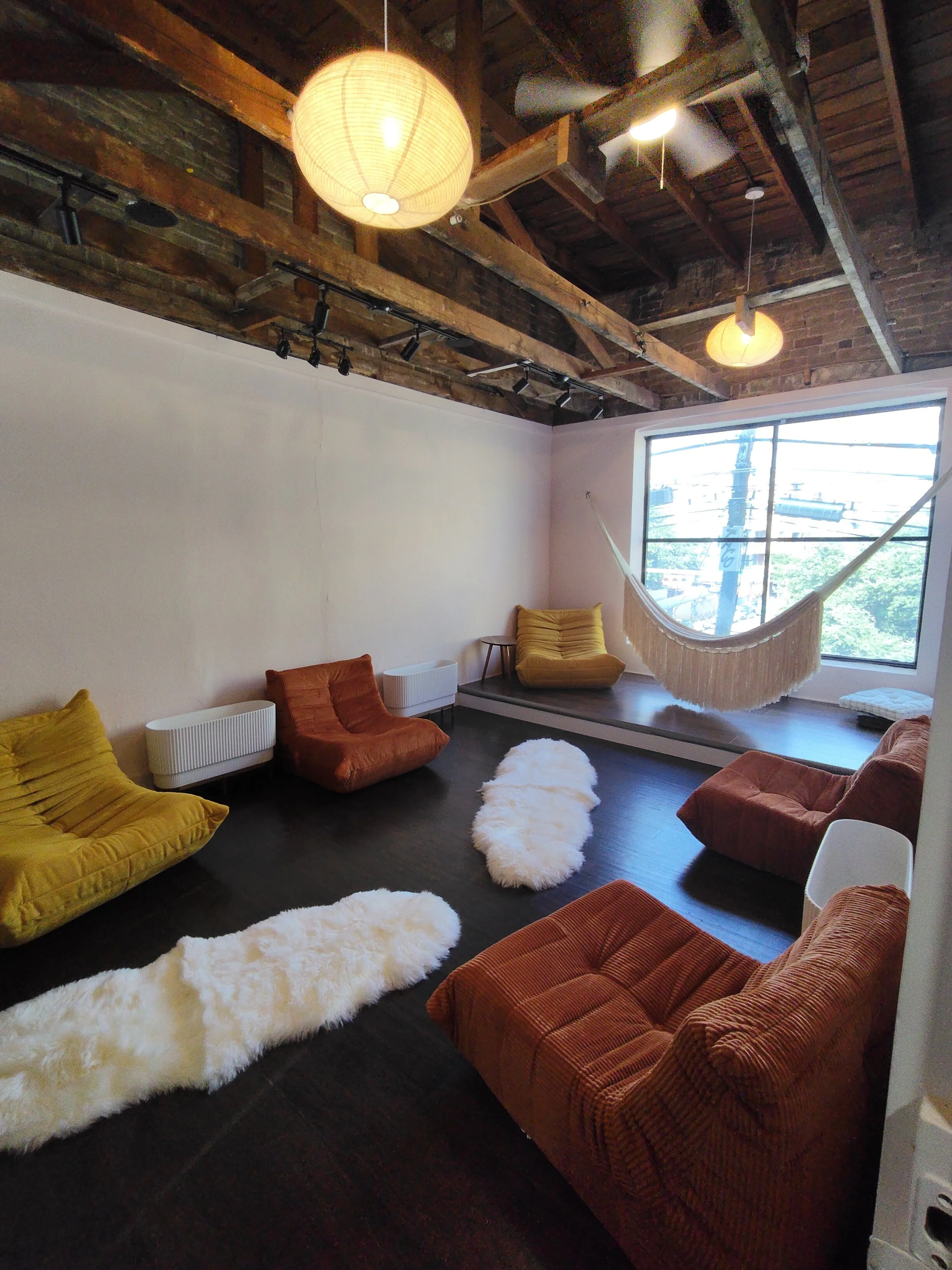 A cozy lounge area with colorful cushions and fluffy white rugs on a dark hardwood floor. The room features an exposed brick and wooden ceiling with hanging pendant lights and track lighting. A large window lets in natural light and offers a view of utility poles and trees outside.