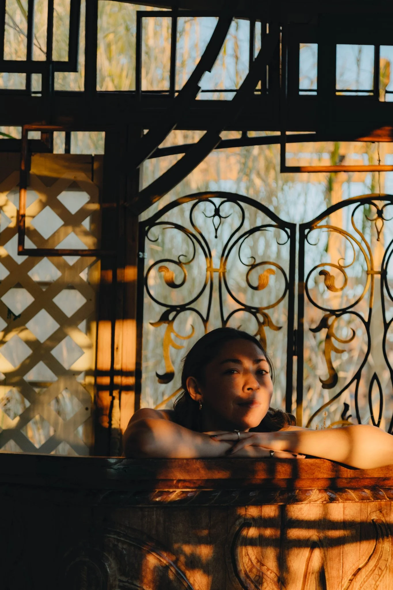A woman relaxing in a wooden hot tub outdoors near a decorative metal gate, with sunlight casting shadows on her face and the surroundings.