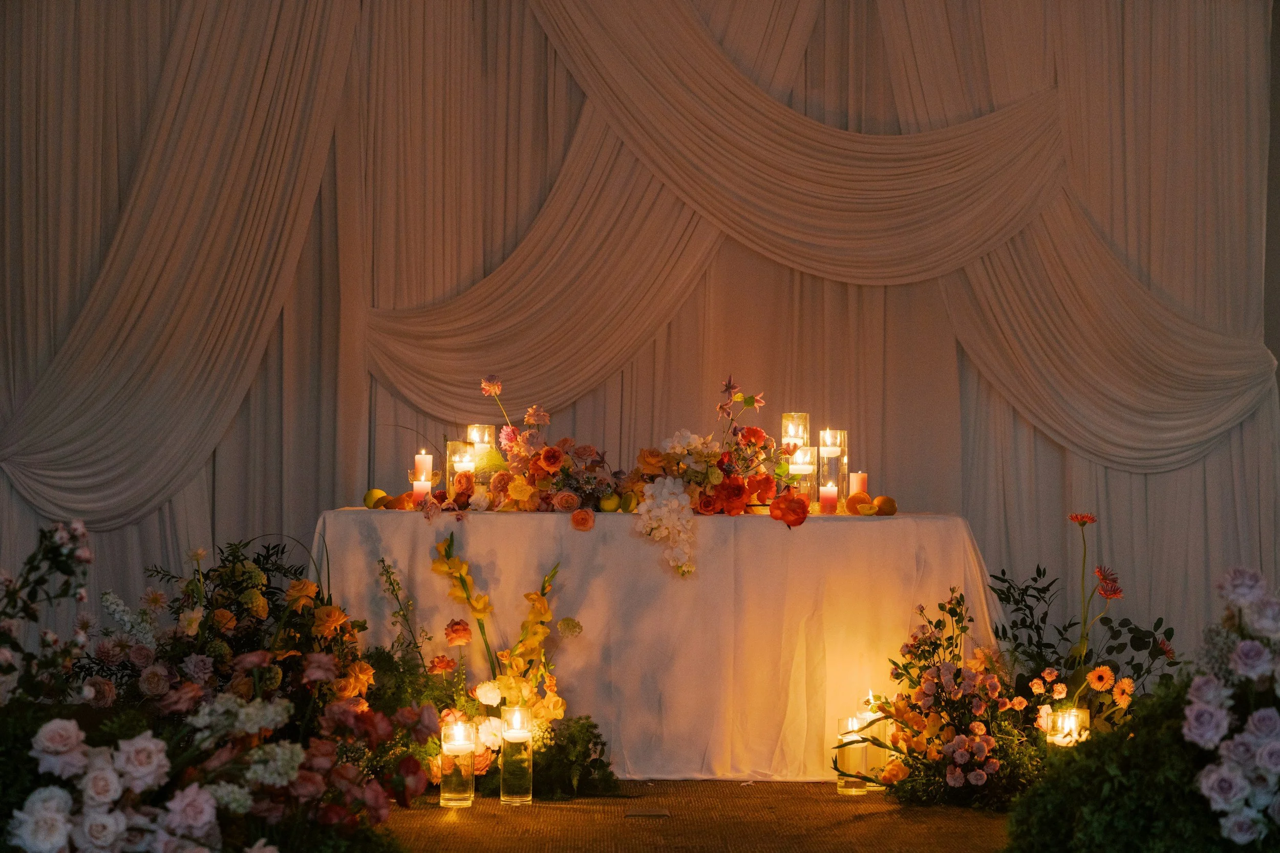 A decorated event table with various candles and colorful flowers, set against draped white fabric.