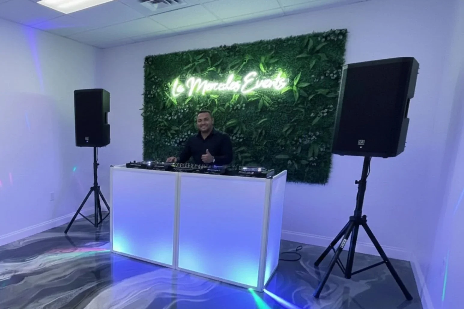 A DJ stands behind a white illuminated DJ booth with a green leafy wall and a neon sign reading 'La Mueble Eventos' in the background. Two large black speakers on stands flank the DJ, and colorful lighting effects are visible on the floor.