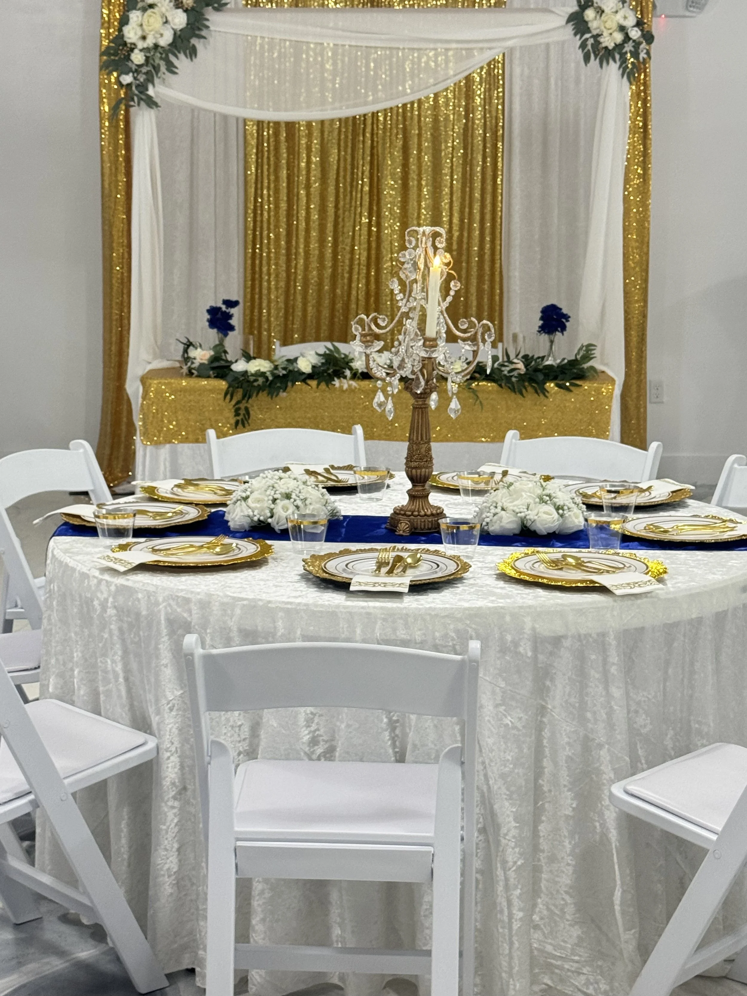 Round banquet table with white tablecloth, set with white and gold plates, gold utensils, and gold napkins. A gold and blue table runner runs down the center, decorated with a floral arrangement of white flowers and a tall bronze chandelier-style cen