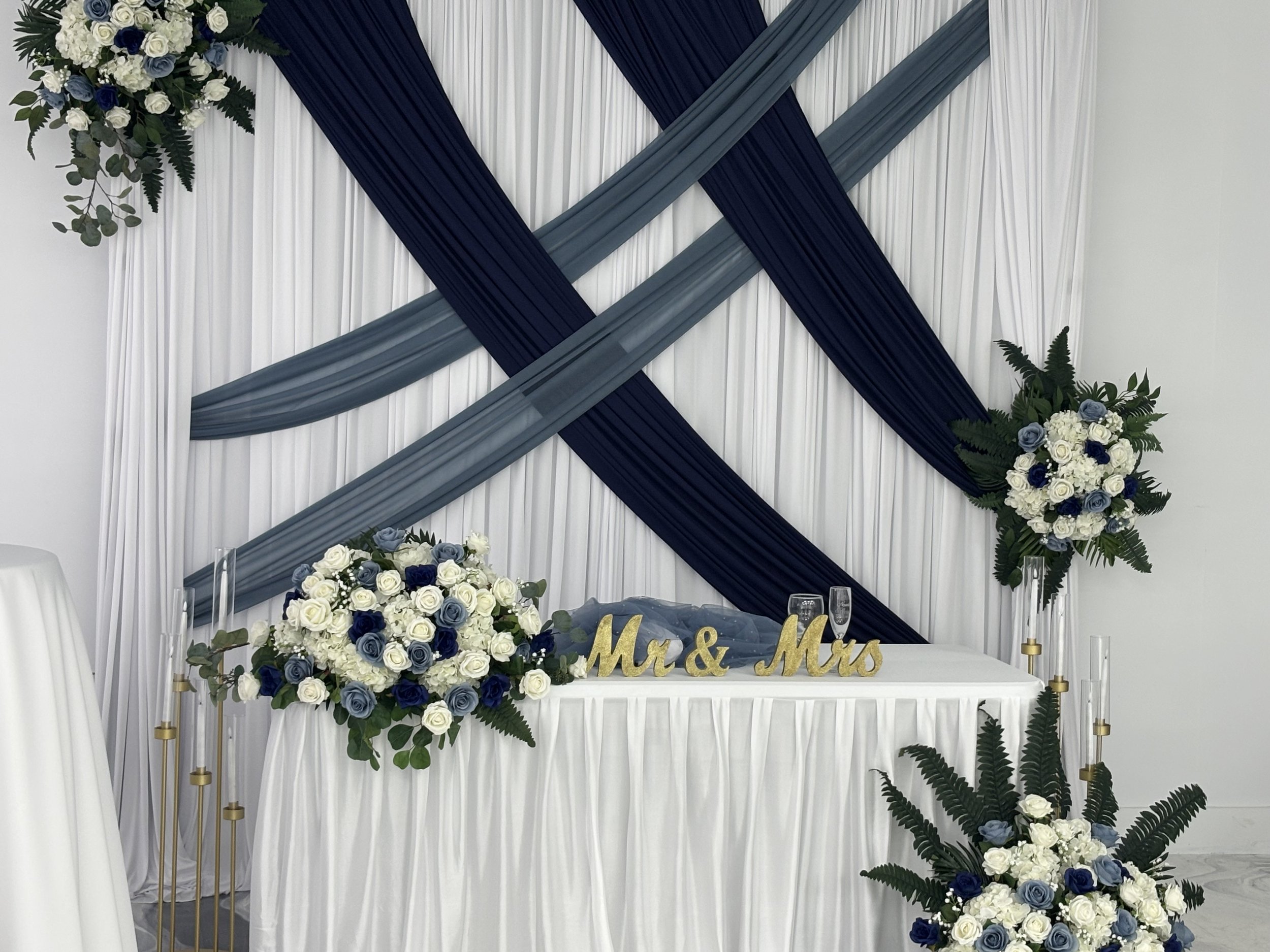 Decorated wedding reception table with floral arrangements in white and blue, gold "Mr & Mrs" sign, and a backdrop with white, navy blue, and gray fabric drapes, flanked by flower arrangements.